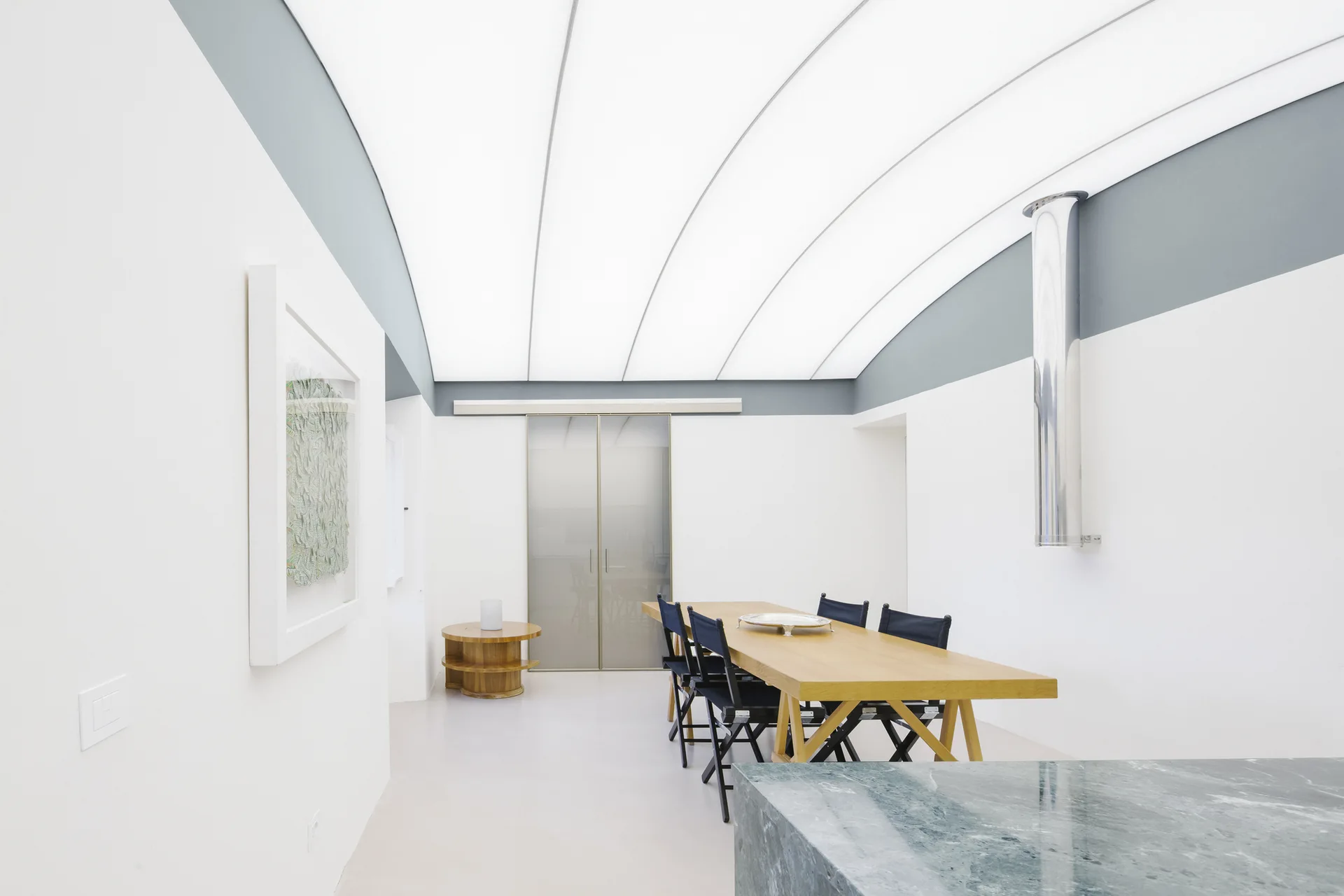 A minimalist dining room featuring a wooden table with black folding chairs beneath a large, illuminated vaulted ceiling and a polished stone kitchen island.