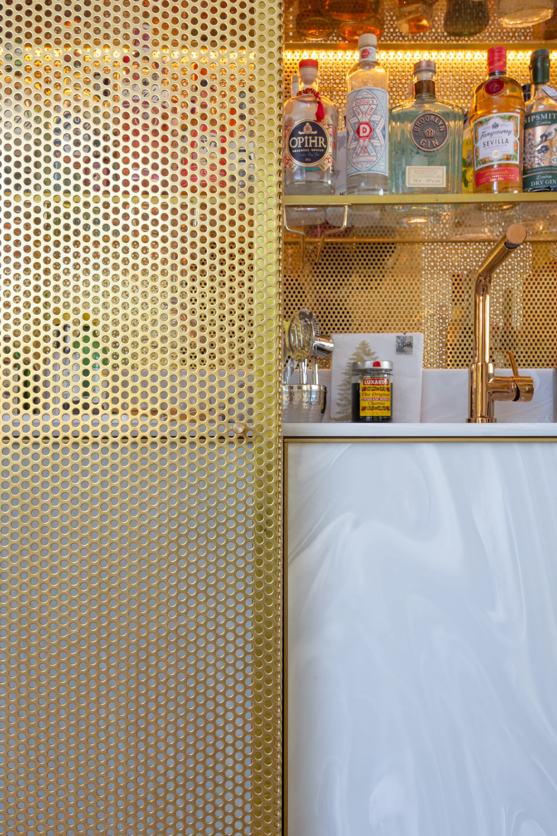 A close-up view of a home bar cabinet featuring a gold perforated metal sliding door, marbled detail, and backlit glass shelves with liquor bottles.