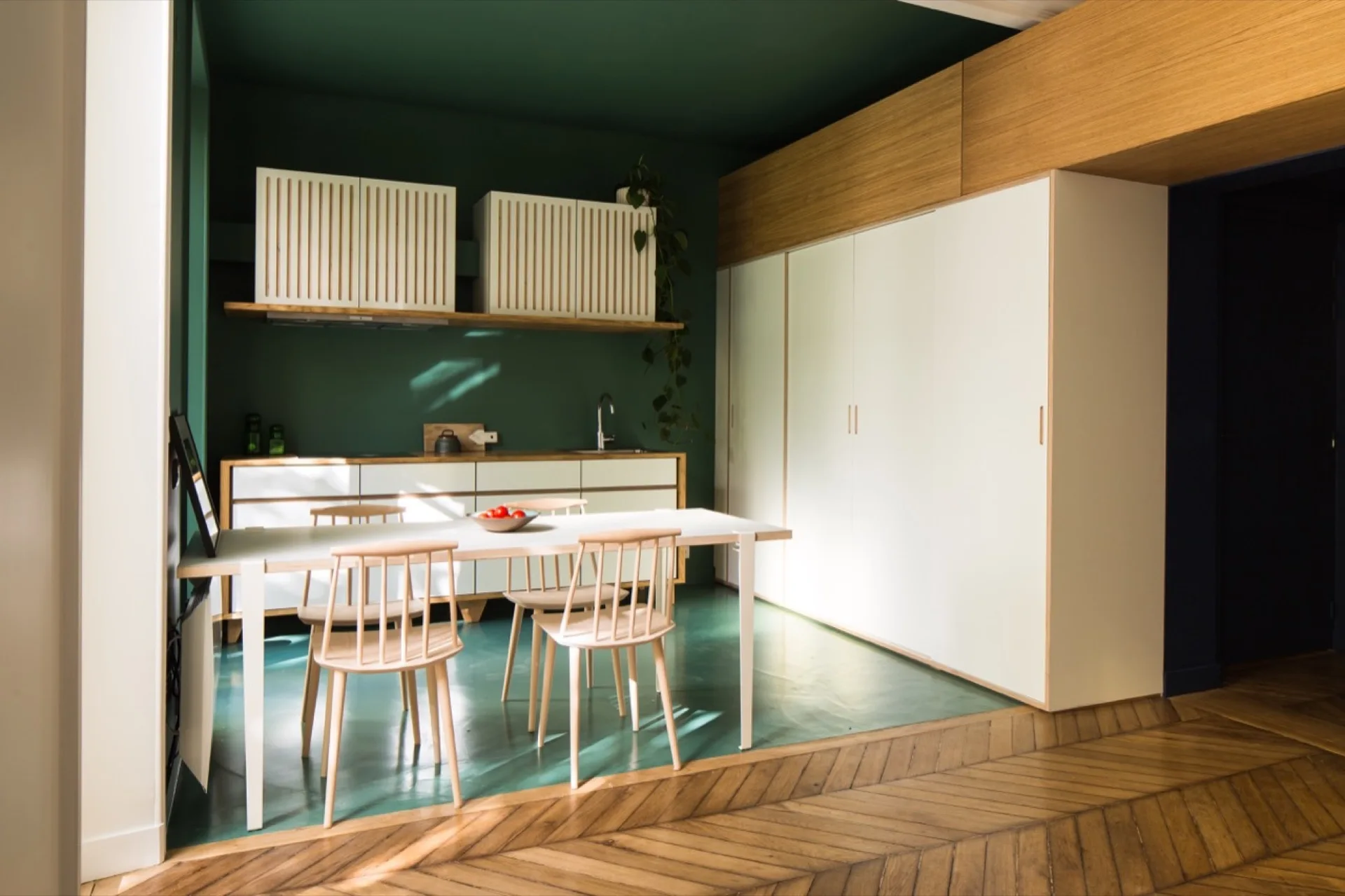A modern kitchen with deep green walls, white cabinets, a light wood dining set, and a step down to a chevron-patterned wood floor.