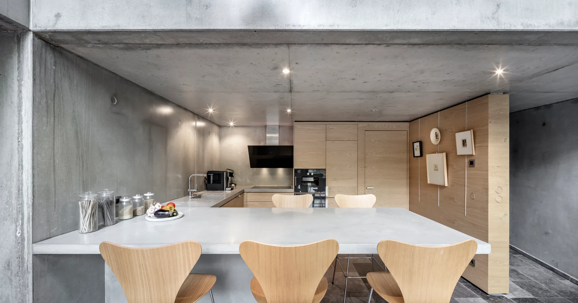 A modern kitchen featuring a concrete island surrounded by light wood chairs, flanked by raw concrete walls and matching oak cabinets.