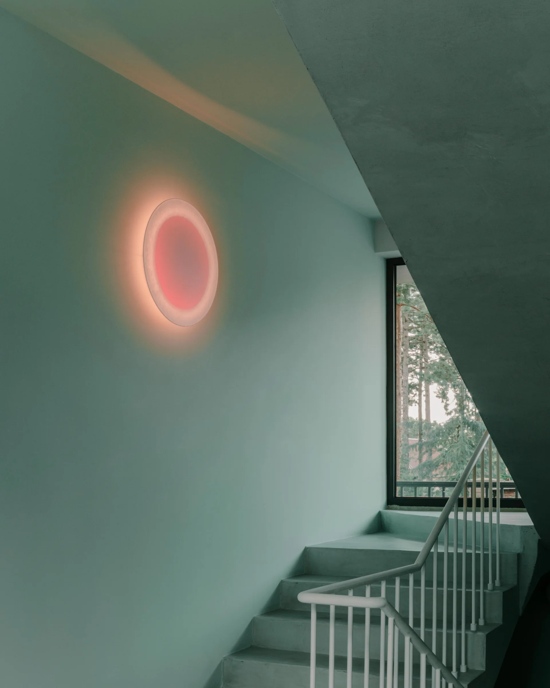 A minimalist concrete staircase with a white metal railing next to a sage green wall featuring a glowing circular red and orange light fixture.
