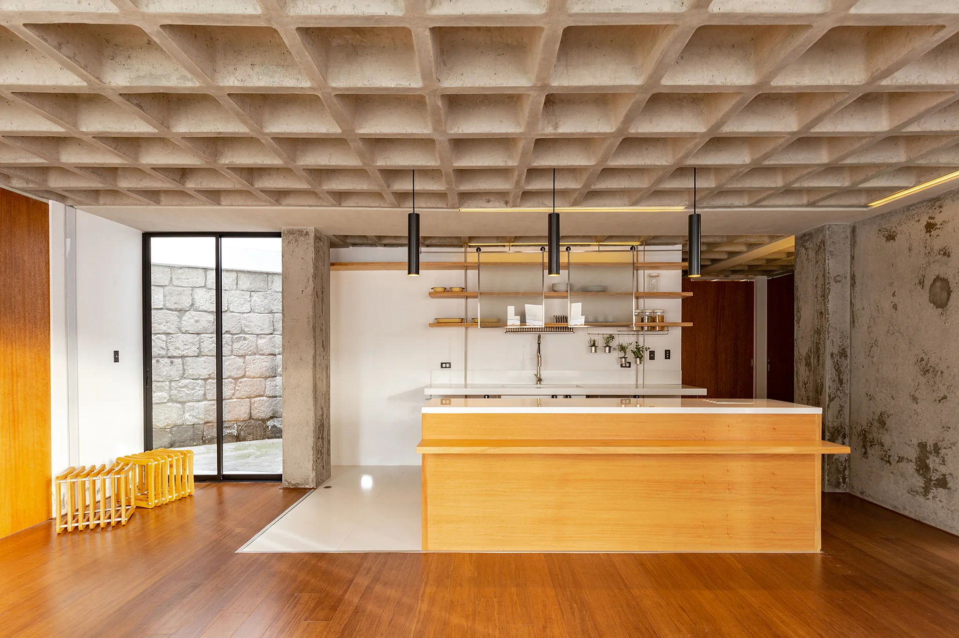A modern kitchen featuring a warm wood island, a concrete waffle slab ceiling, sleek black pendant lights, and open wall shelving.