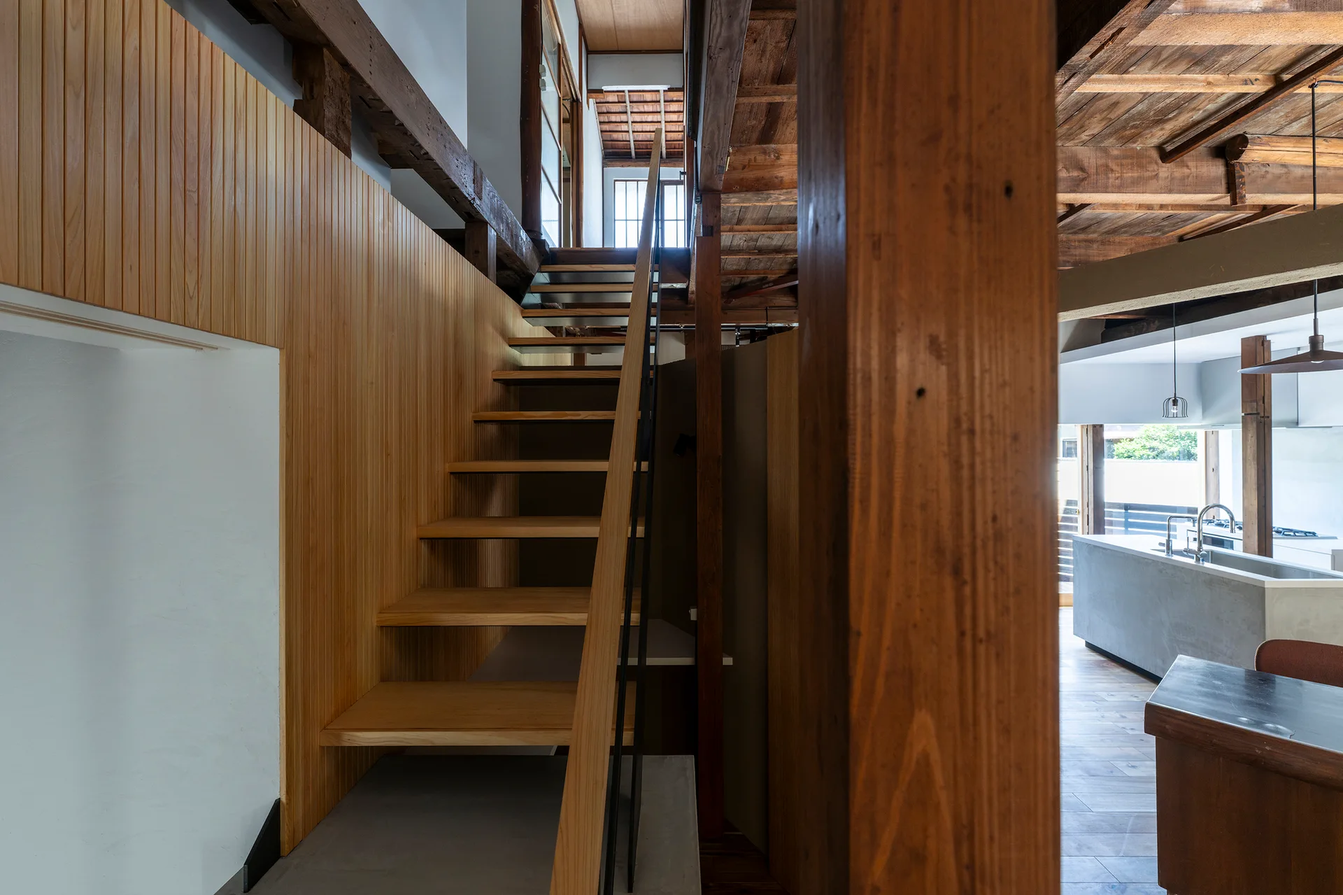 An open wooden staircase with a thin black handrail rises beside a vertical wood slat wall, with a concrete kitchen island visible in the background.