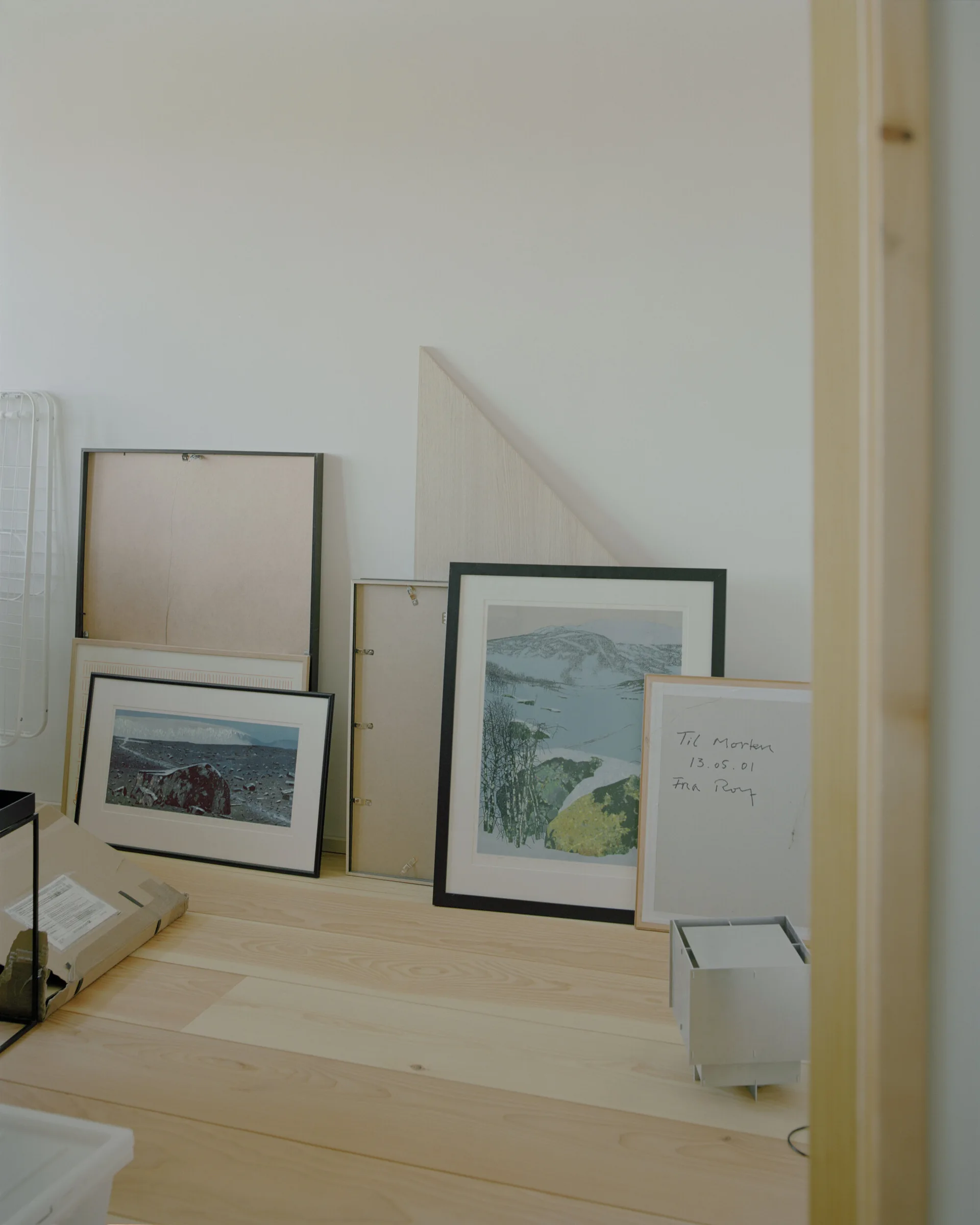 A collection of framed art prints and backing boards lean against a white wall on light-colored wood plank flooring.