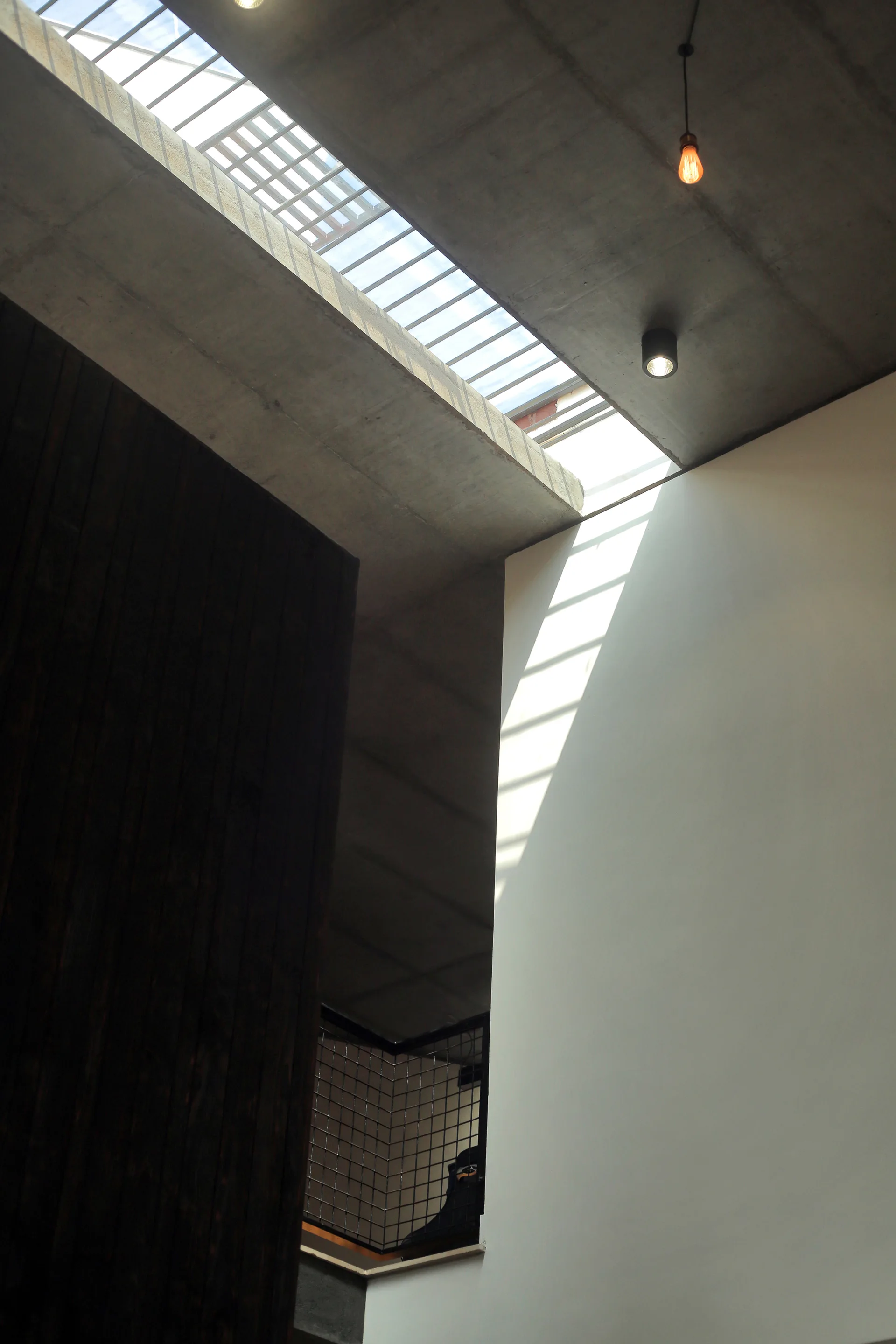 A modern stairwell features an exposed concrete ceiling with a skylight, a dark wood-paneled wall, and a smooth white wall catching sunlight.