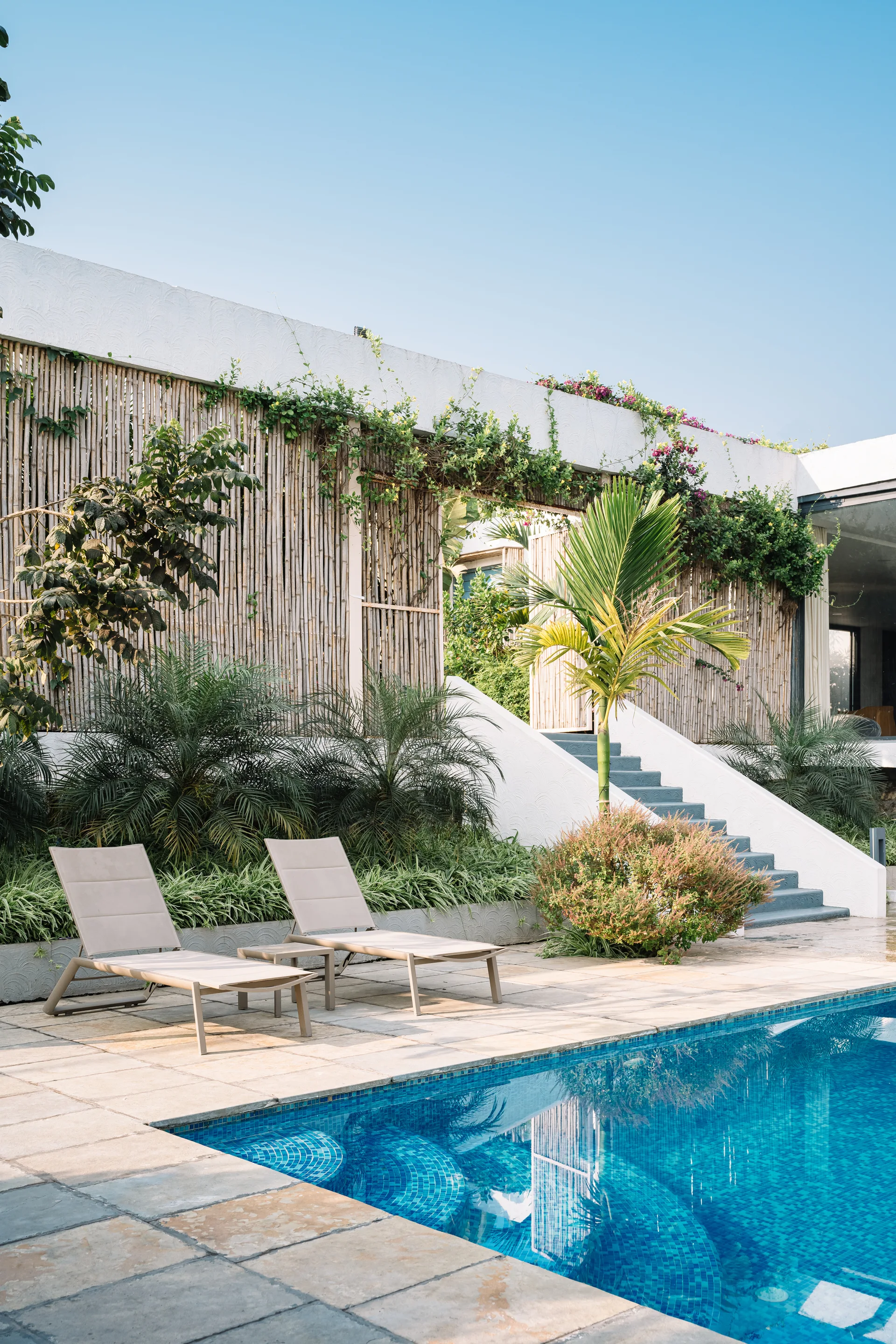 Two beige chaise loungers sit on a stone patio next to a swimming pool, backed by a white staircase and a tall bamboo privacy screen.