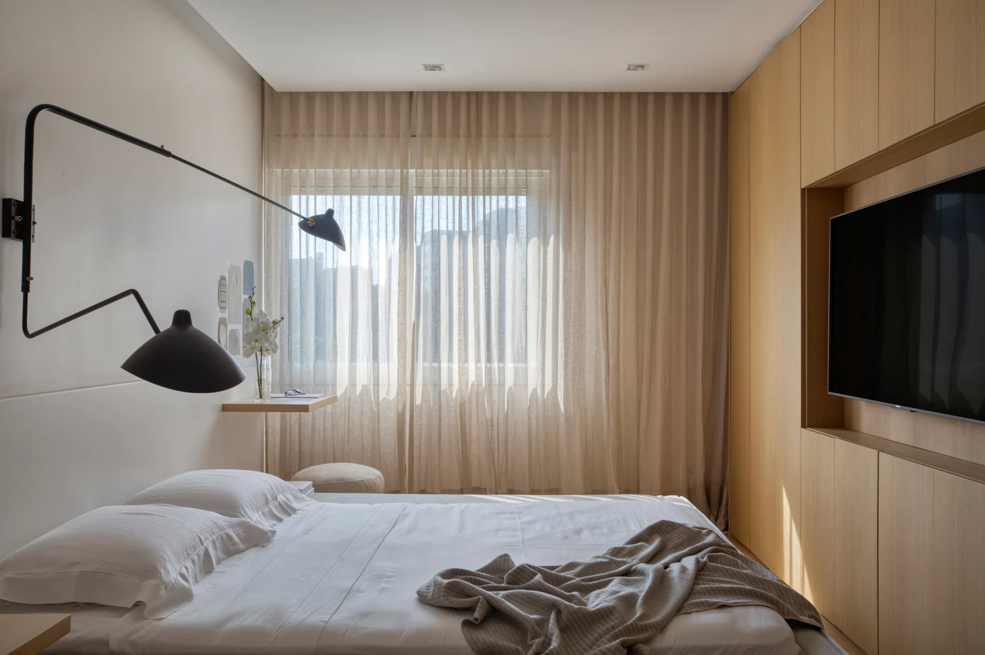 A modern minimalist bedroom featuring a white bed, sheer beige curtains, built-in light wood cabinetry, and a black articulating wall-mounted lamp.