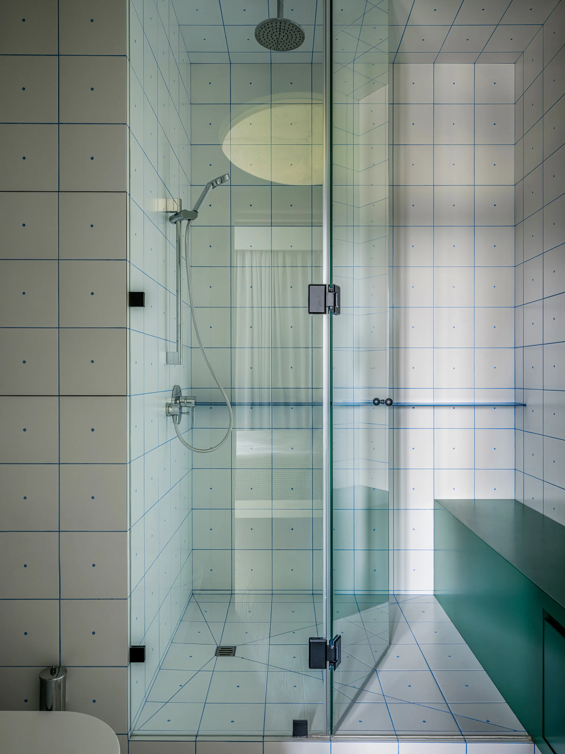 A modern walk-in shower features white ceramic wall tiles with a blue grid and dot pattern, alongside a solid teal built-in bench and a glass door.