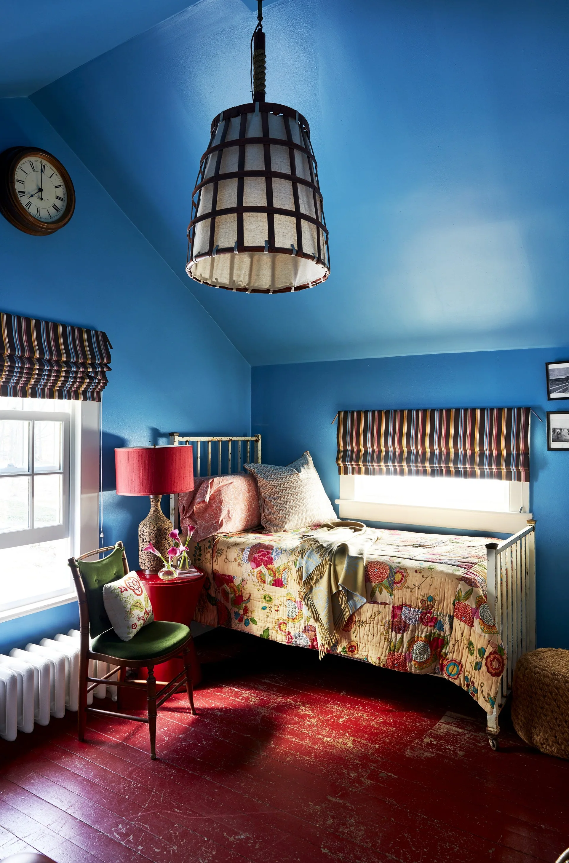 An eclectic bedroom with blue walls, a distressed red wood floor, a metal frame bed with a floral quilt, and a large woven pendant light.