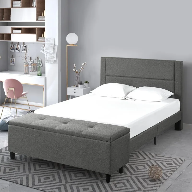A grey upholstered bed with an integrated storage bench at the foot sits on a patterned rug in a bedroom.