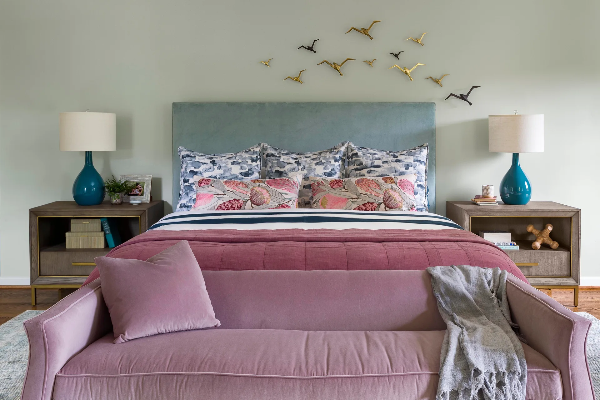 A sage green bedroom features a bed with a blue headboard, pink bedding, a pink upholstered bench, and gold bird sculptures on the wall.