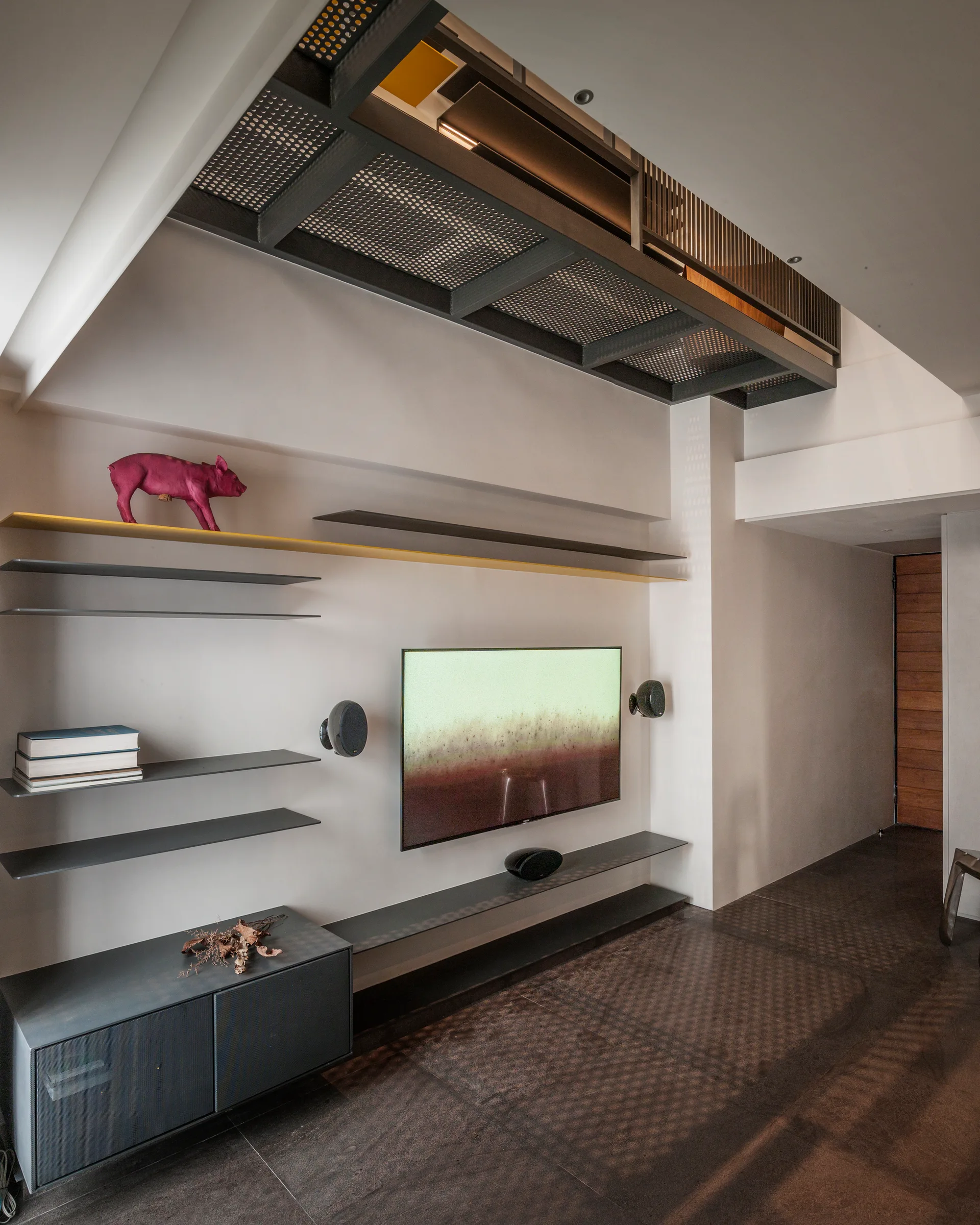 A wall-mounted television is surrounded by minimalist metal shelving below a perforated industrial steel ceiling structure.