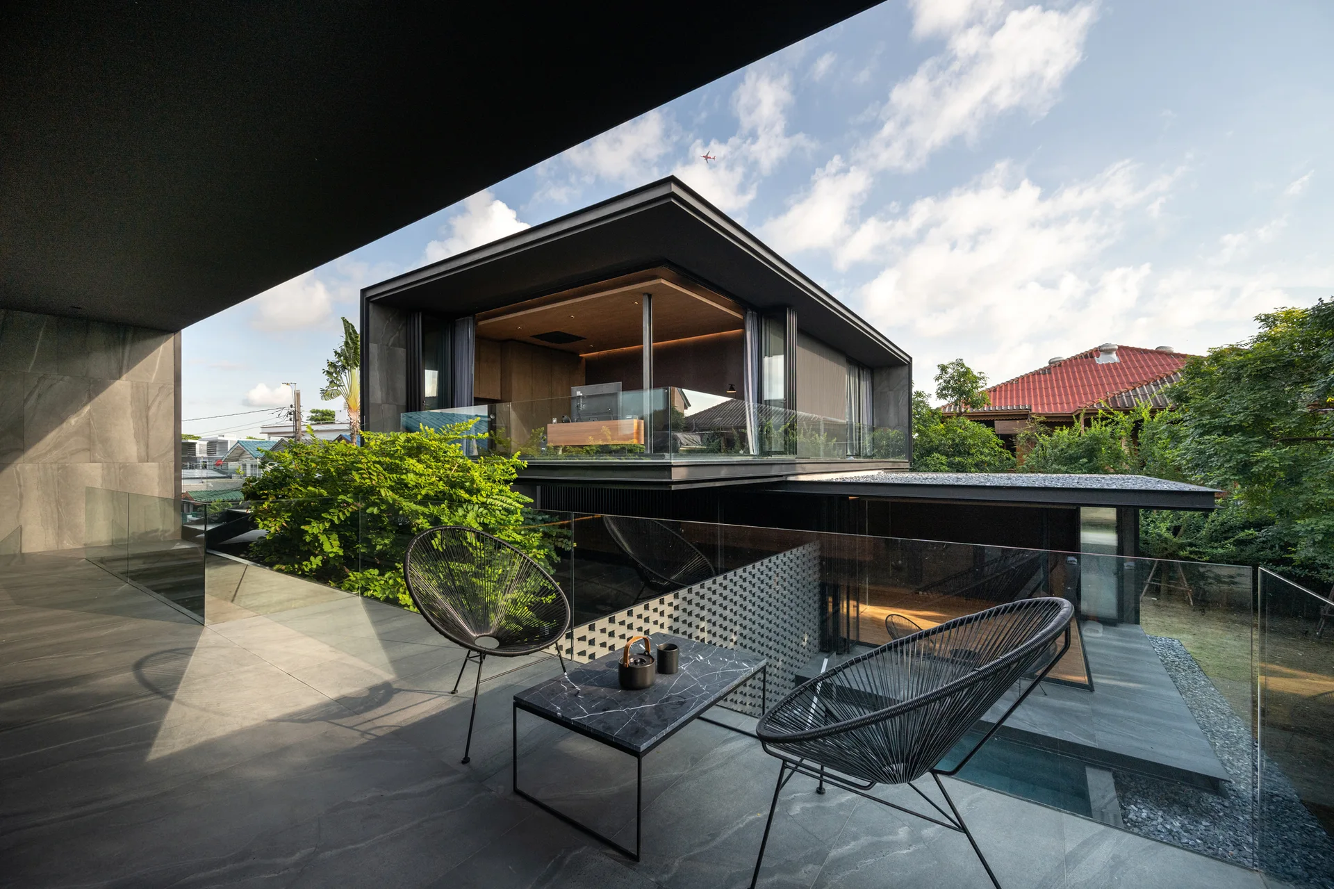 Two black wire-frame chairs and a small marble table sit on a spacious grey stone tile terrace with a glass railing overlooking a modern home.