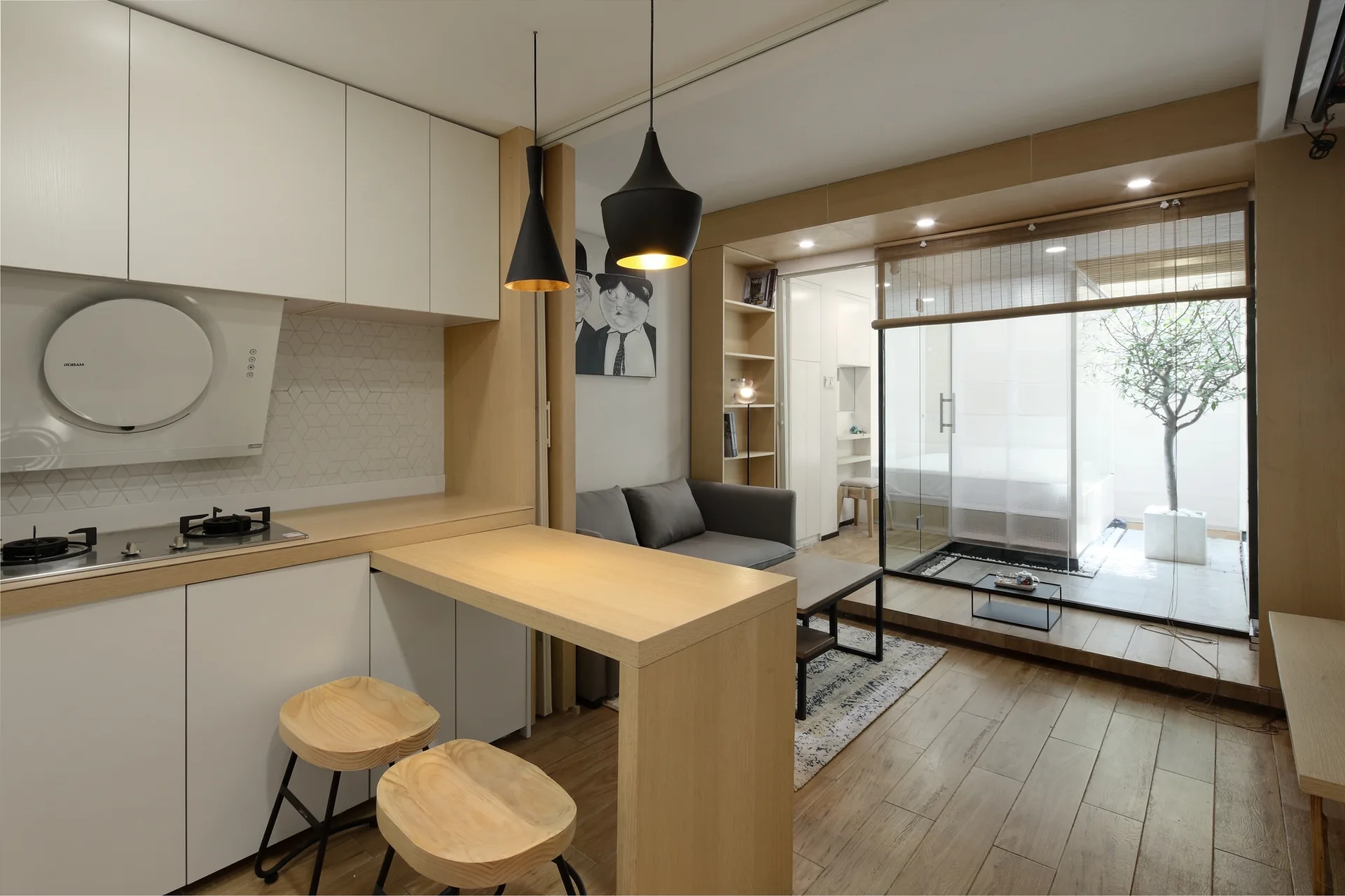 A compact modern kitchen with white cabinets, a light wood peninsula, and wooden stools next to a small living area with a sliding glass terrace door.