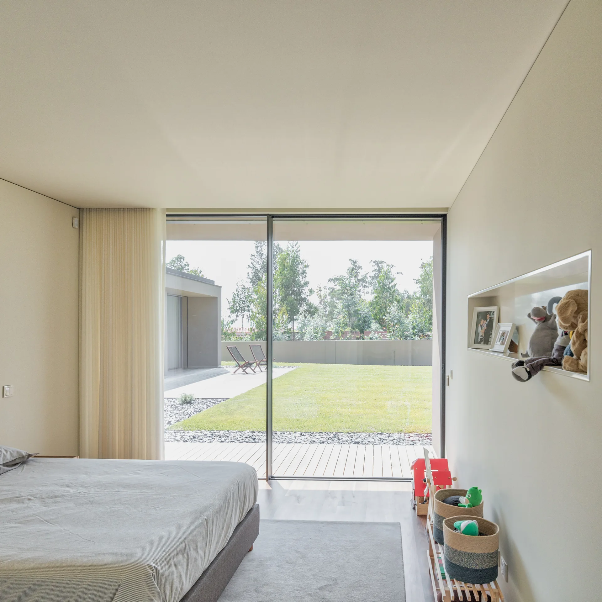 A minimalist bedroom features a low platform bed, a recessed wall shelf with toys, and a large sliding glass door opening to a green lawn.