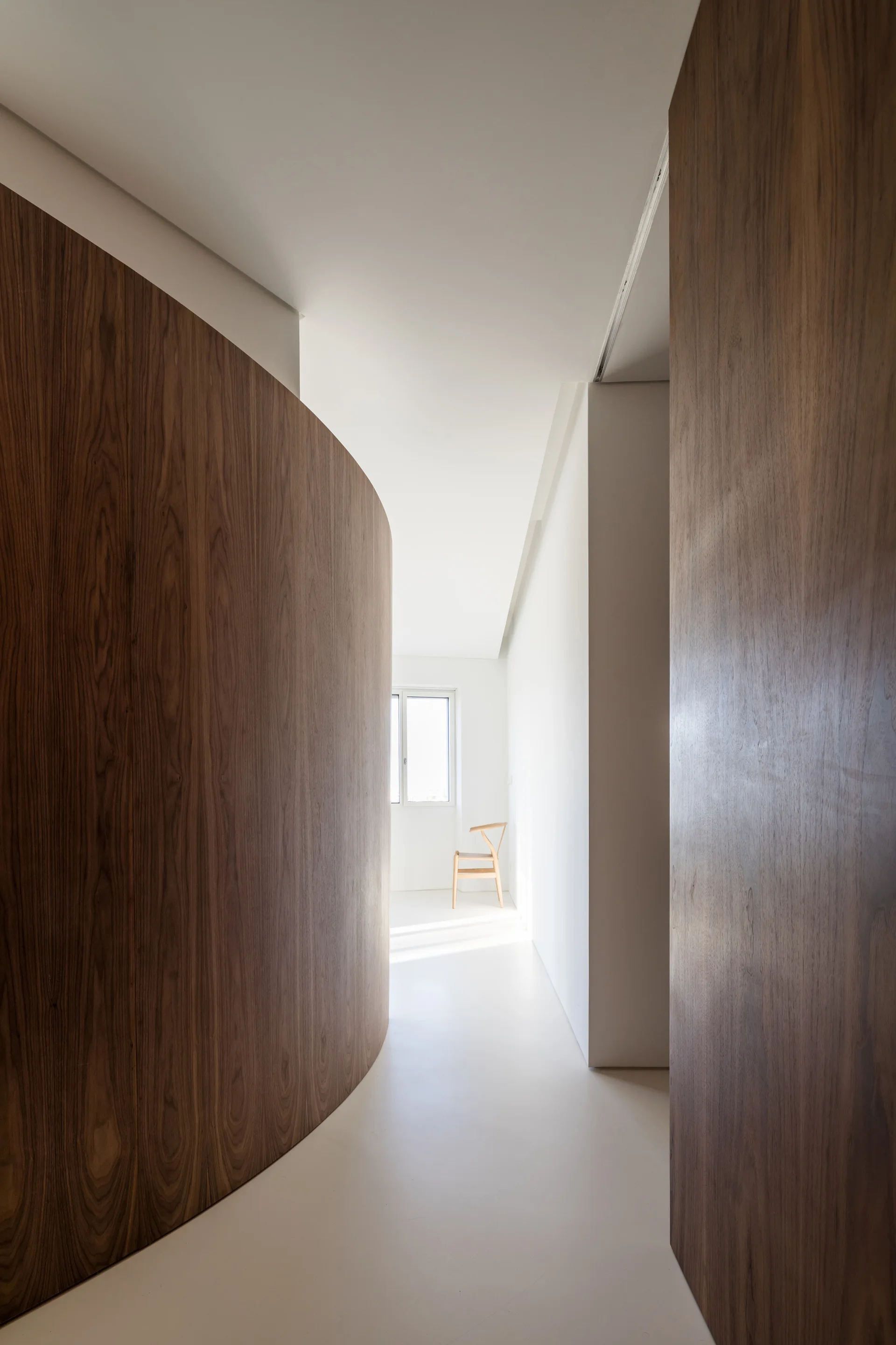 A minimalist white hallway features a prominent curved wood-paneled wall leading to a window and a single wooden chair.