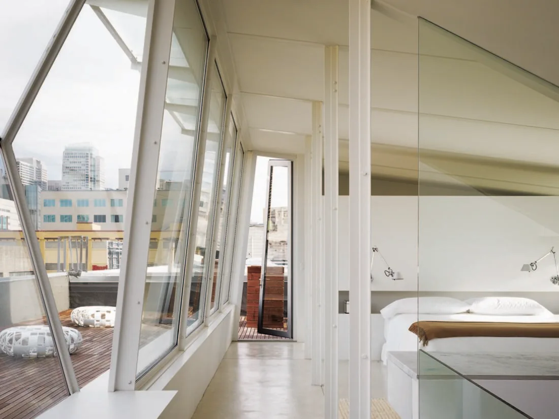 A minimalist bedroom features an angled glass wall looking out onto a wooden deck with city views, next to a bed with white bedding.