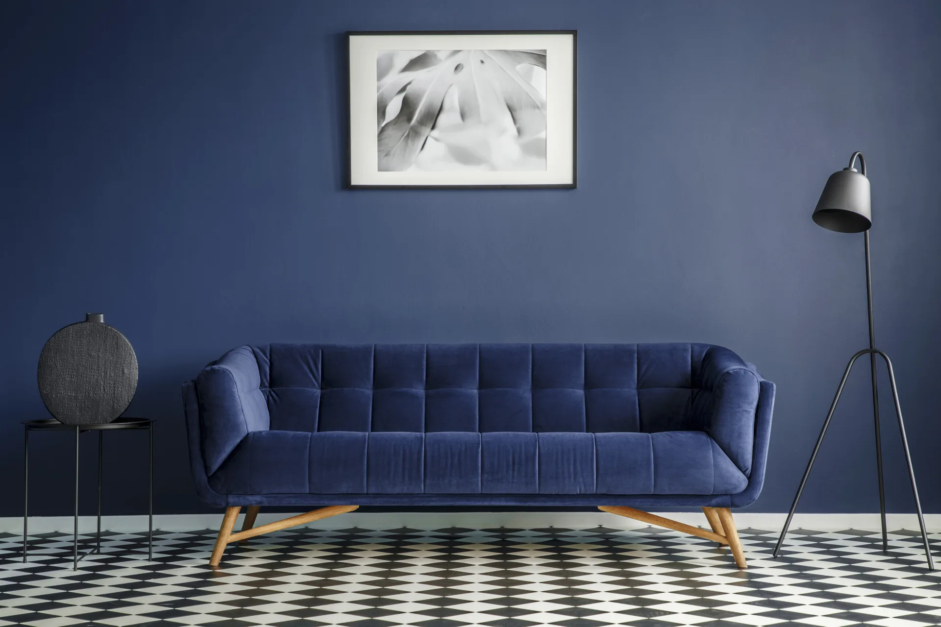 A mid-century modern blue velvet sofa sits against a navy blue wall on a black and white checkered floor flanked by a tripod lamp.