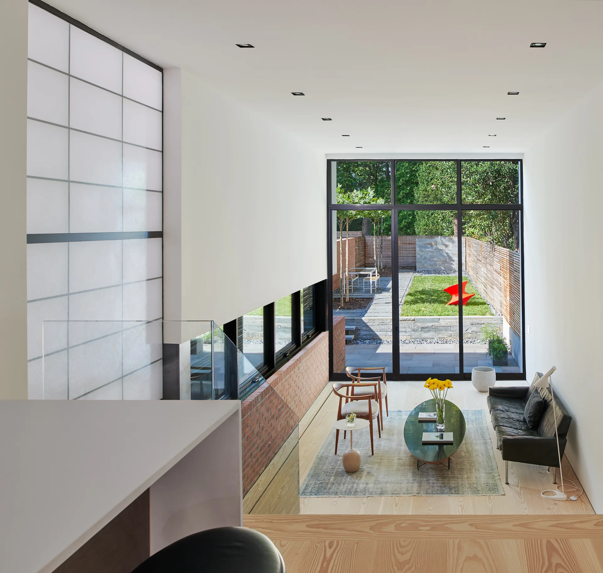 A sunken modern living room with a black leather sofa, mid-century wood armchairs, and floor-to-ceiling glass doors opening to a backyard garden.