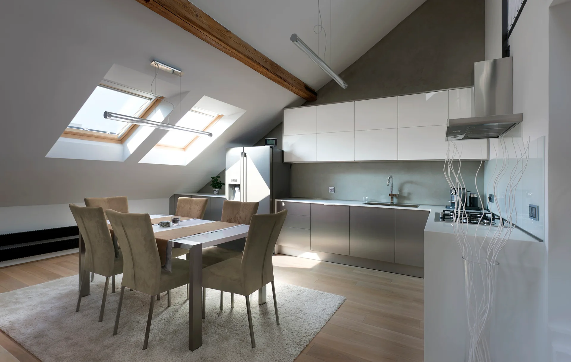 A modern attic kitchen features white and grey flat-panel cabinets under a sloped ceiling with skylights next to a dining table with chairs.