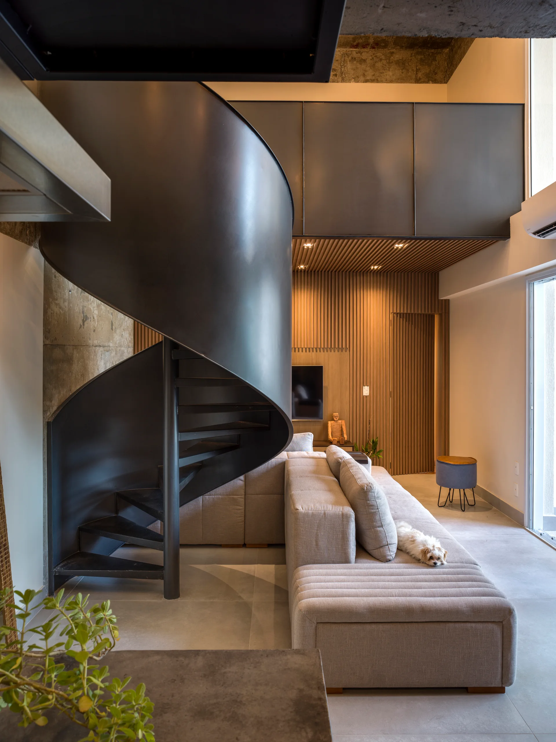 A black metal spiral staircase stands in a modern living room next to a grey sectional sofa and a vertical wood slatted feature wall.