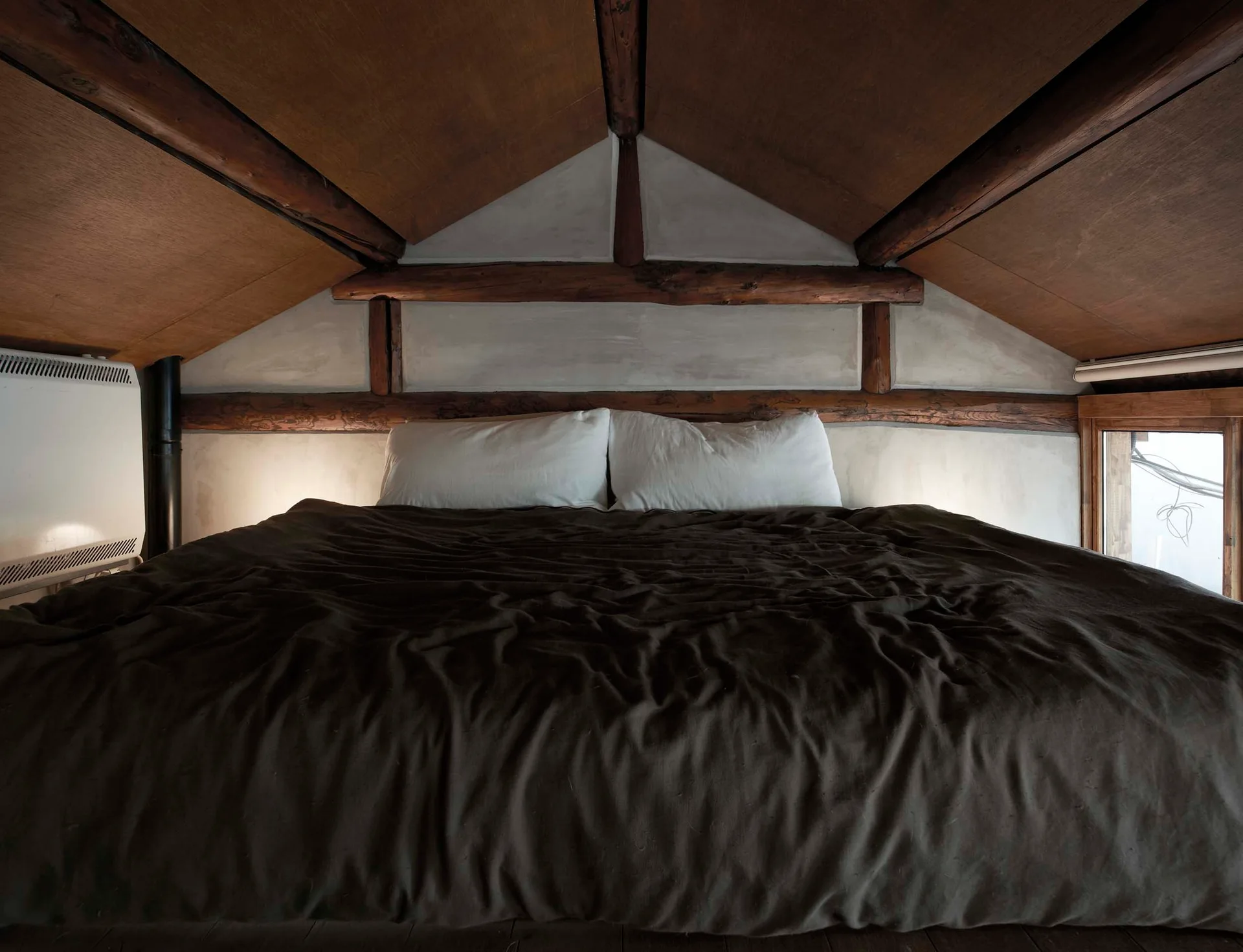 An attic bedroom with a low wood-paneled ceiling, exposed timber beams, and a bed with dark brown bedding and white pillows.