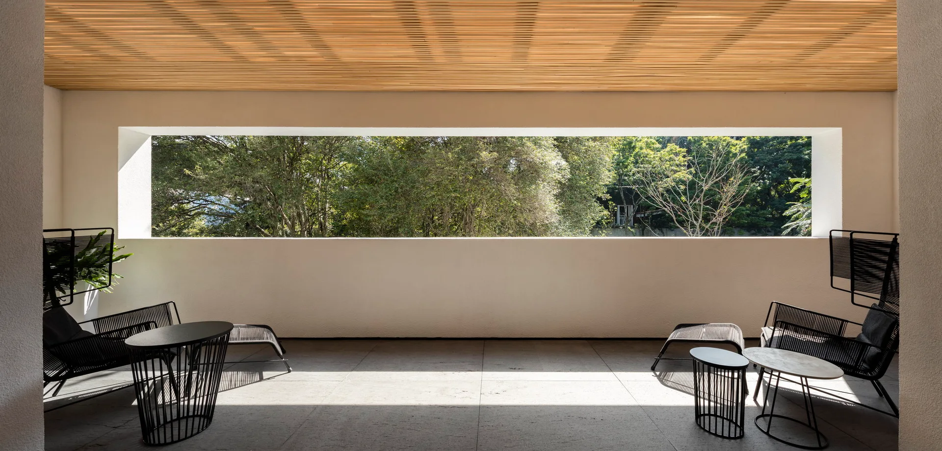 A minimalist outdoor terrace featuring black metal patio furniture under a slatted wood ceiling with a wide rectangular opening showing green trees.