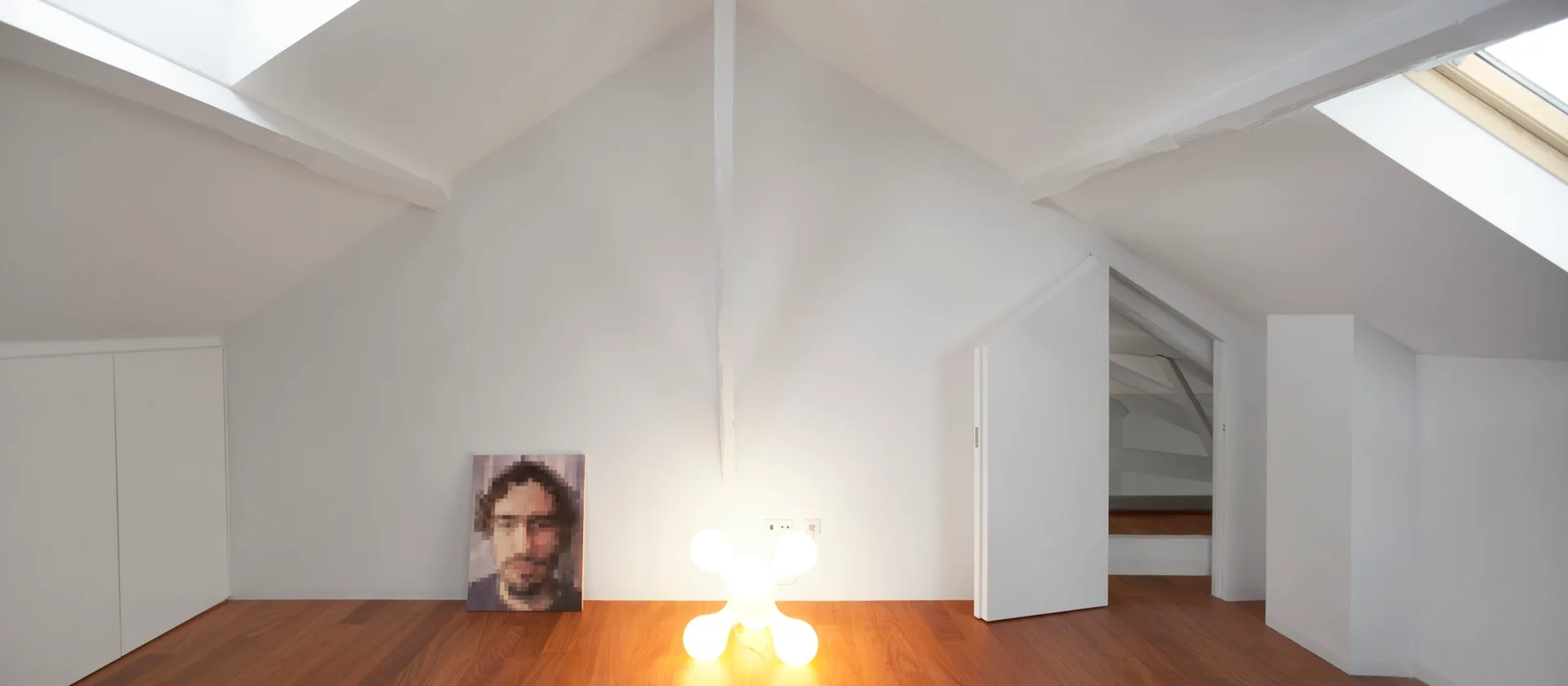 A white attic room with sloped vaulted ceilings, skylights, warm hardwood floors, a sculptural floor lamp, and artwork resting against the wall.