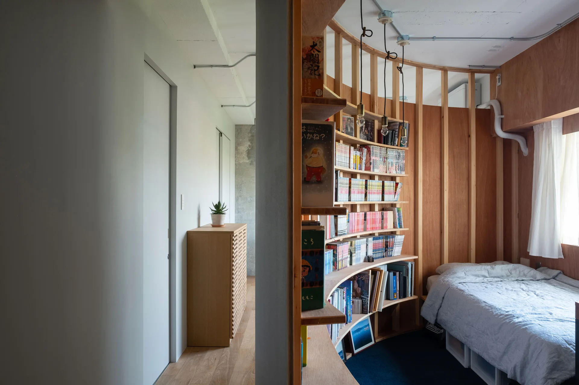 A compact bedroom features a low bed next to a curved plywood bookshelf partition wall, with a hallway visible on the left.