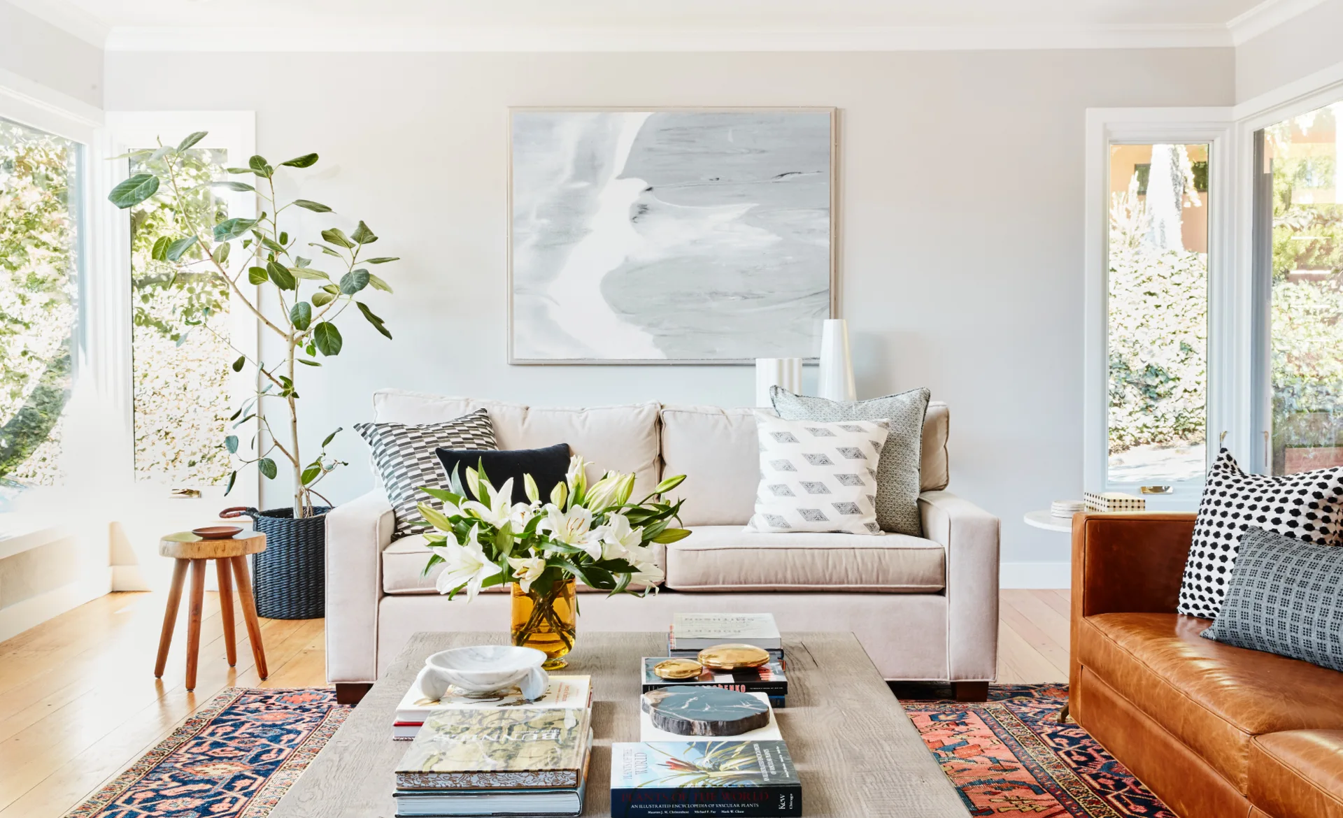 A bright living room features a beige fabric sofa, a brown leather armchair, and a wooden coffee table styled with books on a traditional patterned rug.