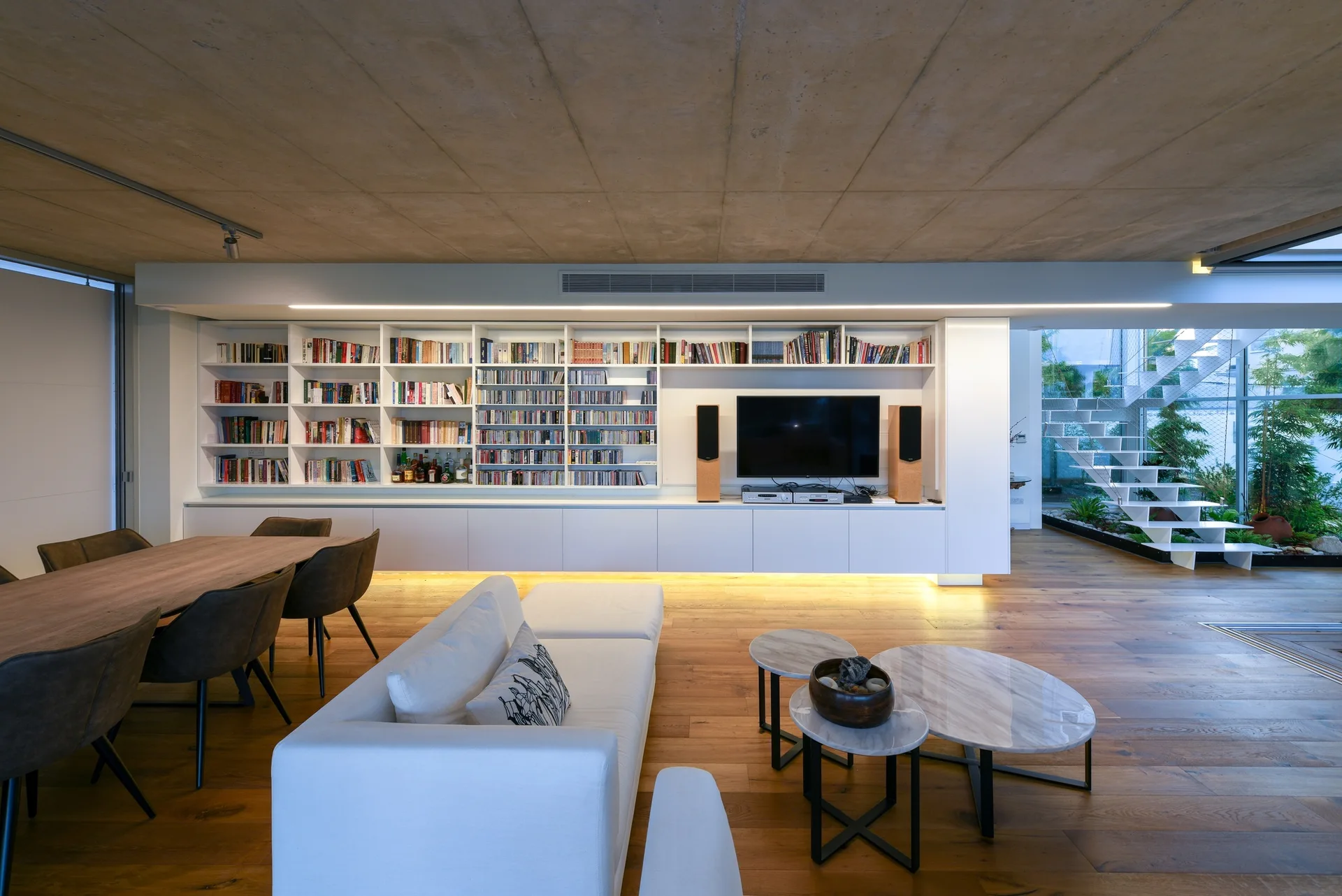 A contemporary living room features an exposed concrete ceiling, a white built-in bookshelf with integrated lighting, and a white sofa.