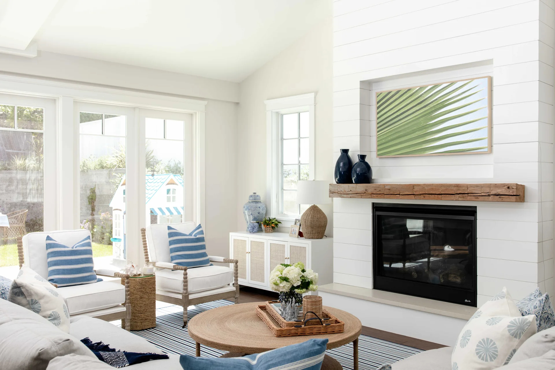 A bright coastal living room featuring a white shiplap fireplace with a rustic wood mantel, white armchairs, and a round woven coffee table on a striped rug.