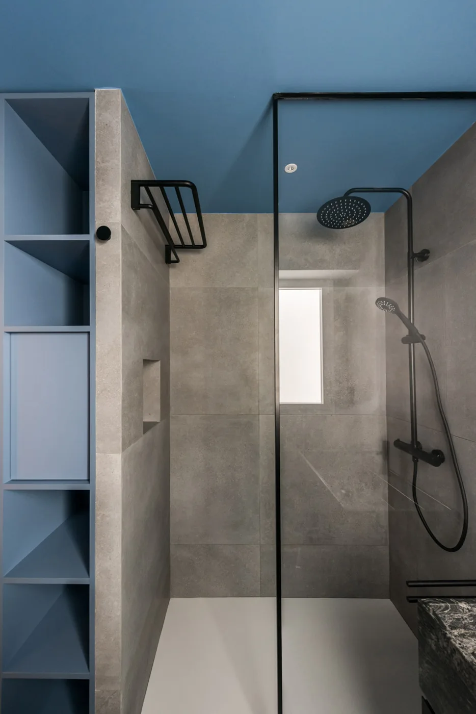 A modern bathroom features a glass-enclosed shower with grey tiles, matte black fixtures, a blue ceiling, and matching blue built-in shelving.