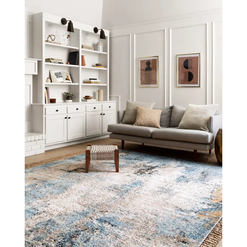 A gray sofa sits on a large blue patterned area rug next to white built-in bookshelves with black sconces in a bright living room.