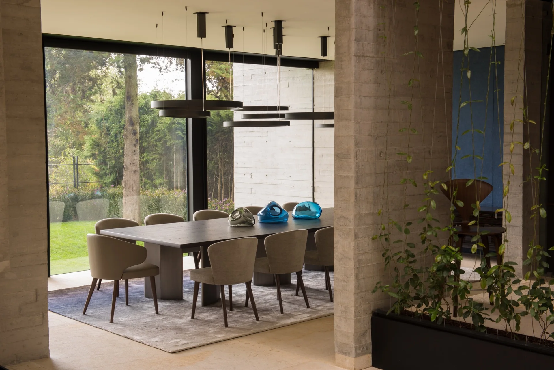 A dark wood dining table with beige upholstered chairs sits on a gray rug between a concrete column and large windows showing a green garden.