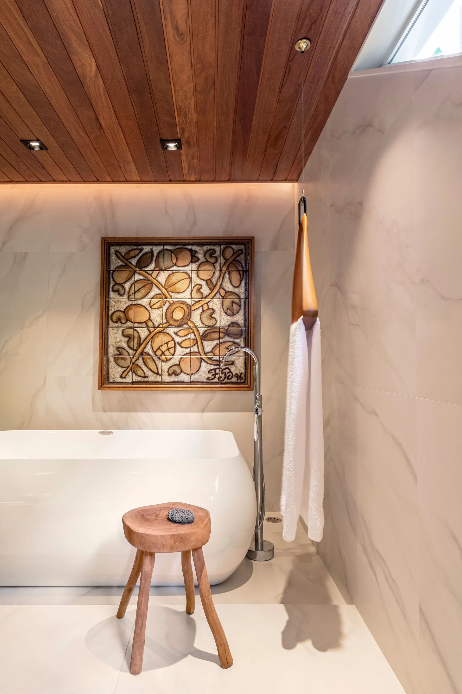 A white freestanding bathtub sits below a wood plank ceiling and large-format marble-look tiled walls, paired with a rustic wooden tripod stool.