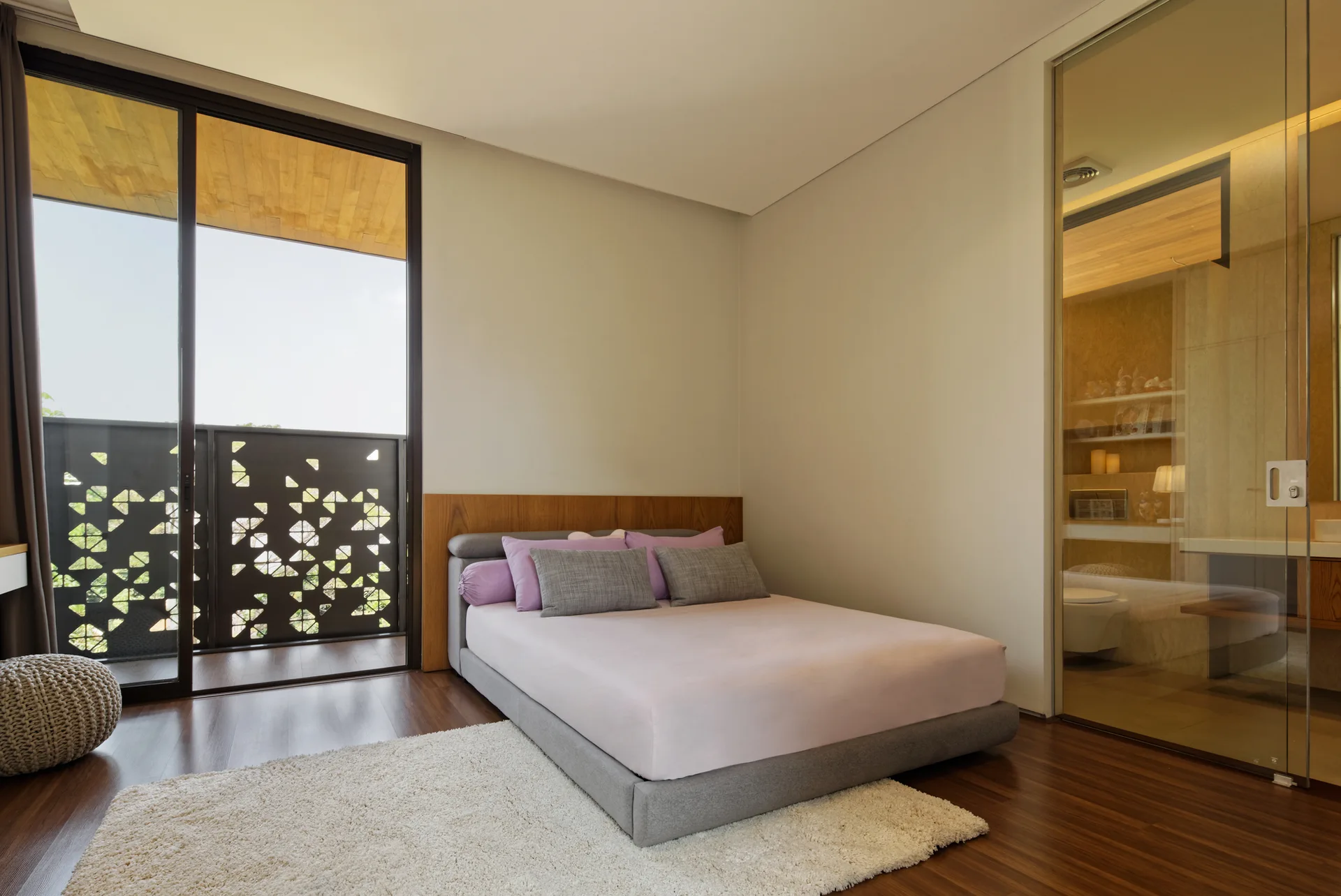 A contemporary bedroom features a low grey platform bed, a textured shag rug, a sliding balcony door, and a glass bathroom partition.