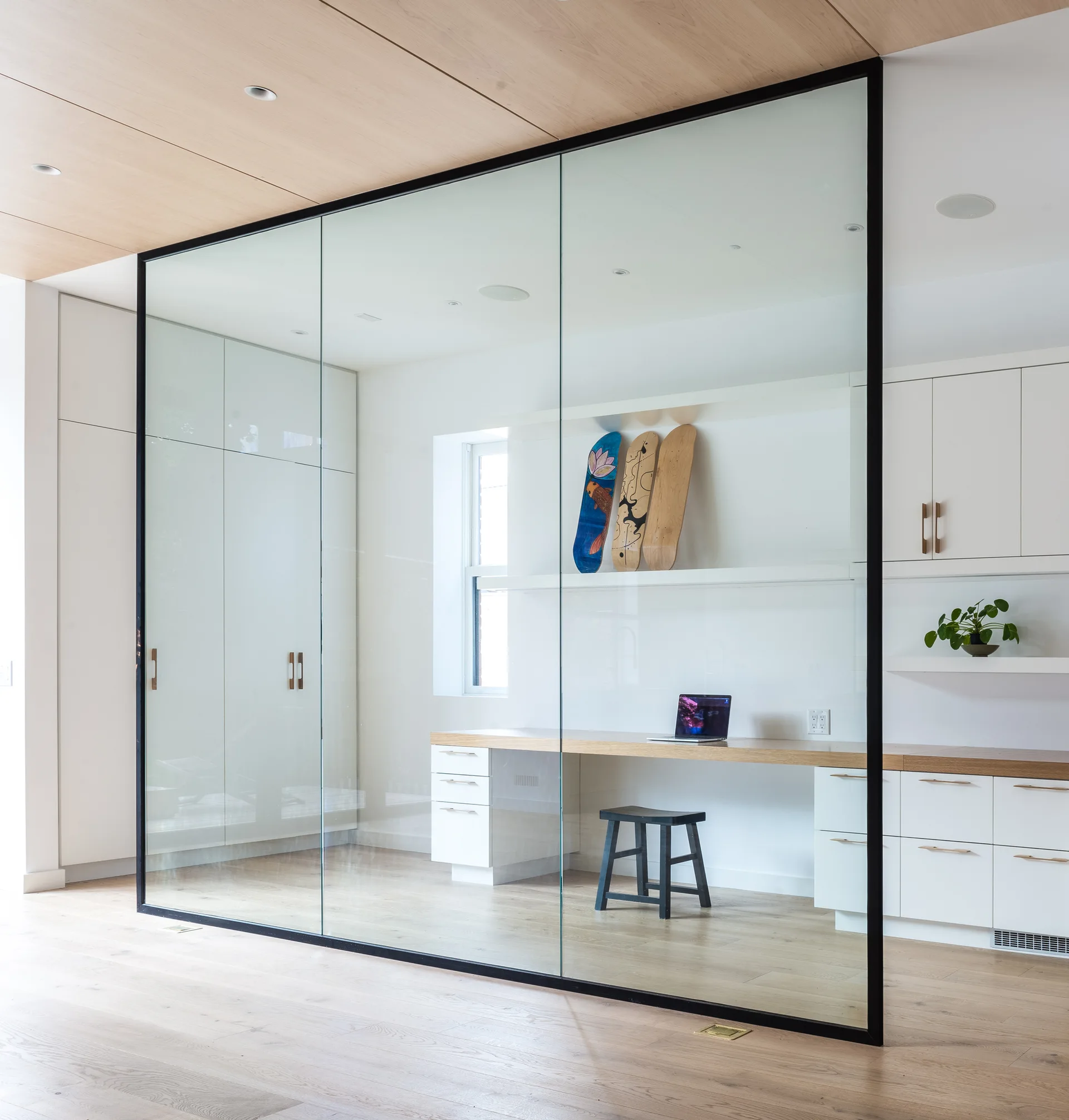 A modern home office with a built-in wooden desk and white cabinetry is enclosed by a floor-to-ceiling glass partition with a black metal frame.