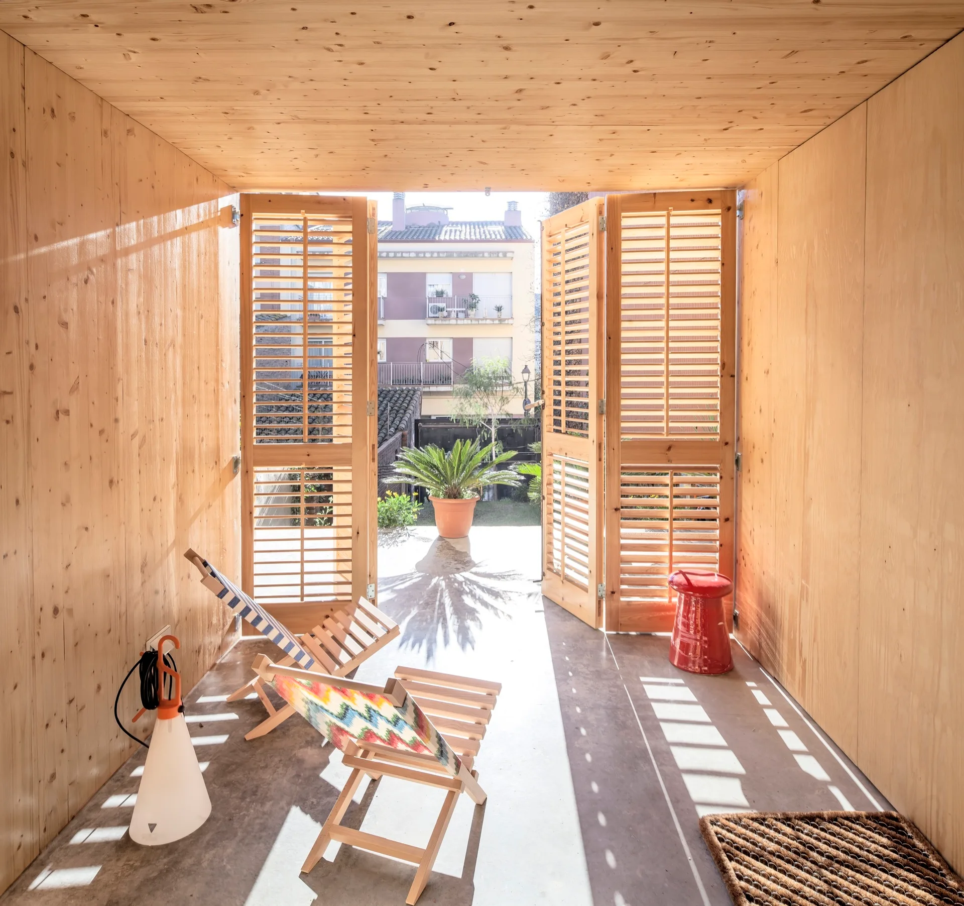 A minimalist room with light wood paneled walls, a concrete floor, folding lounge chairs, and open wooden louvered doors leading outside.