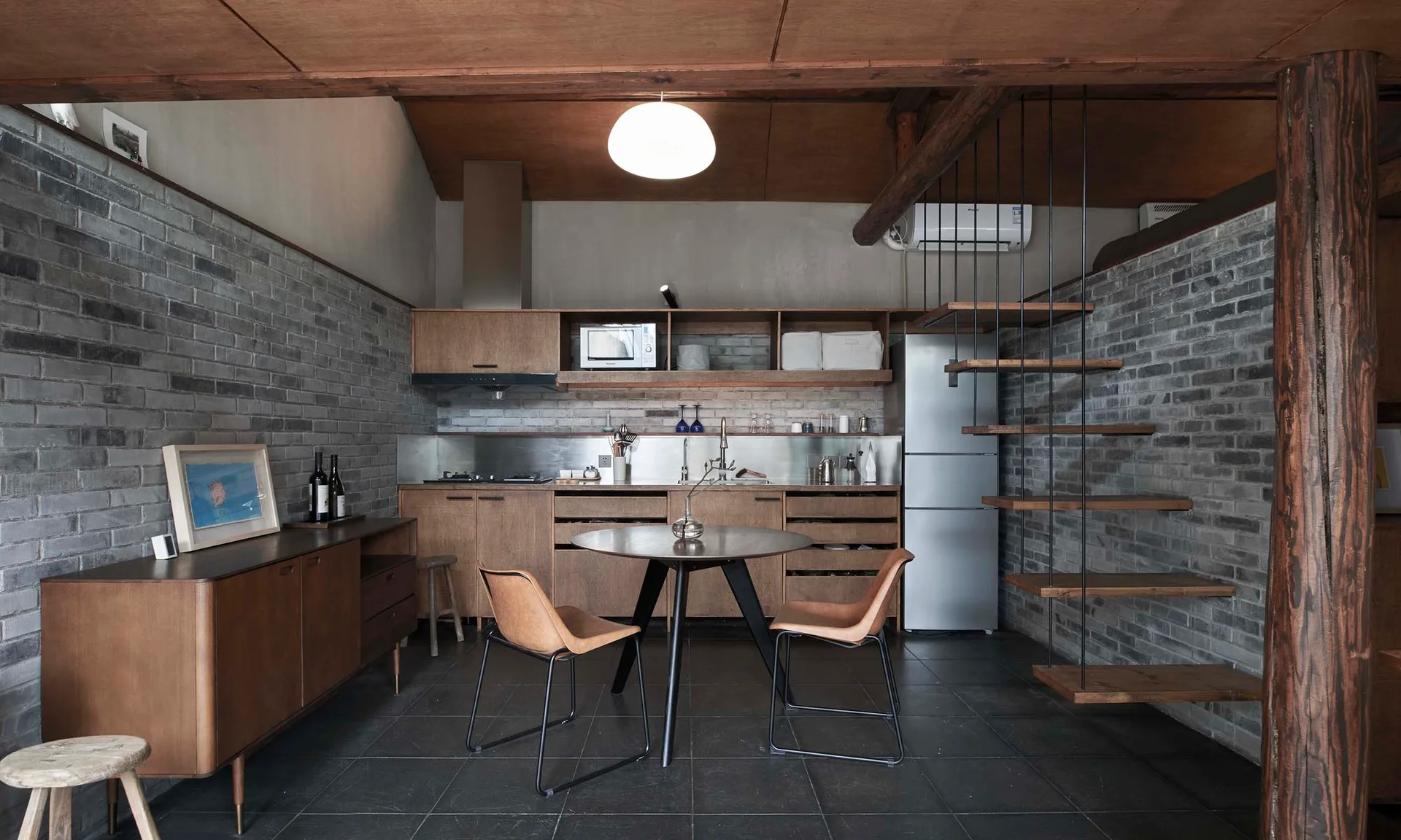 An industrial kitchen features grey brick walls, wood cabinets, a round central dining table with tan leather chairs, and floating stairs.