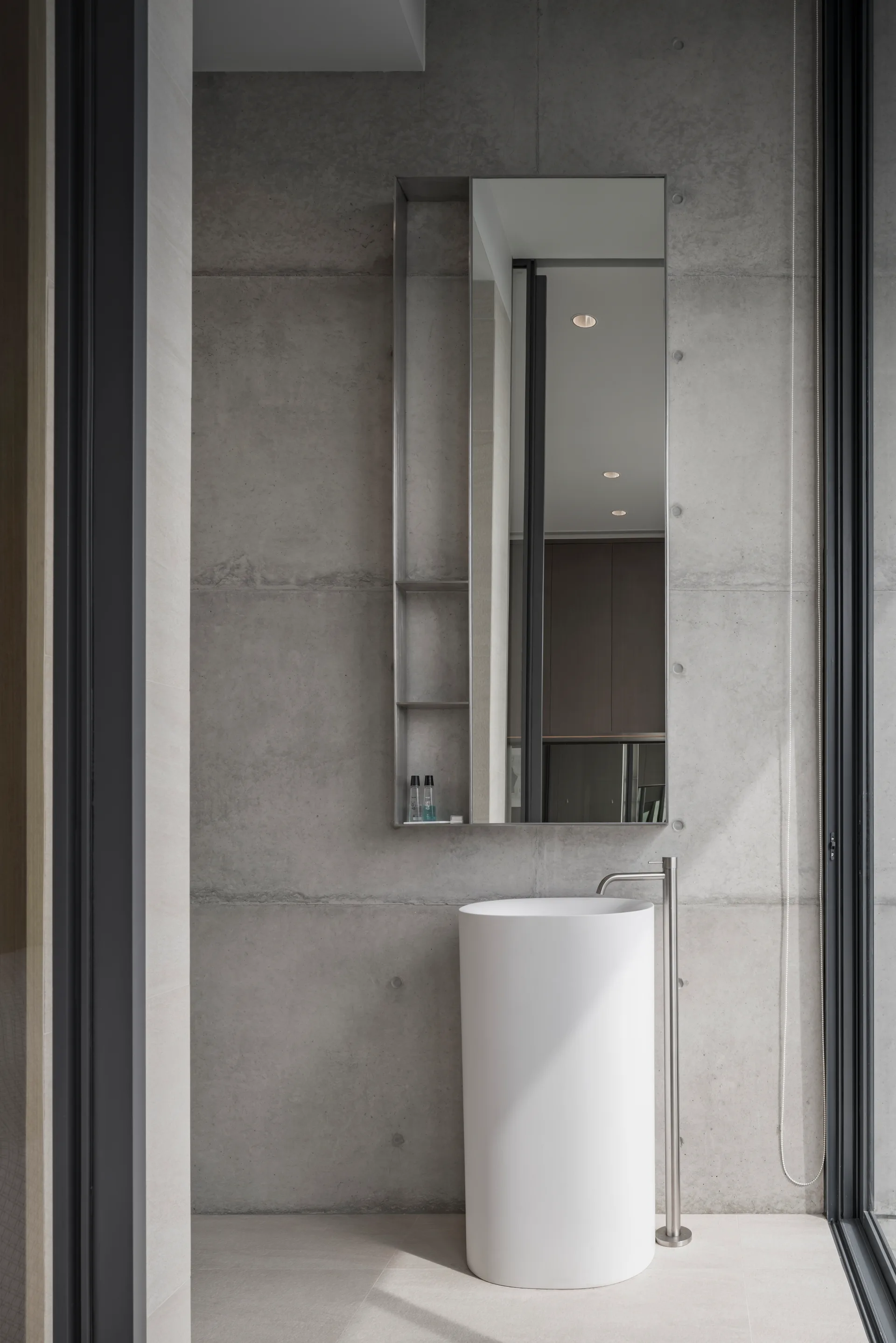 A minimalist bathroom featuring a cylindrical white pedestal sink, a floor-mounted metal faucet, and a mirror against a raw concrete wall.