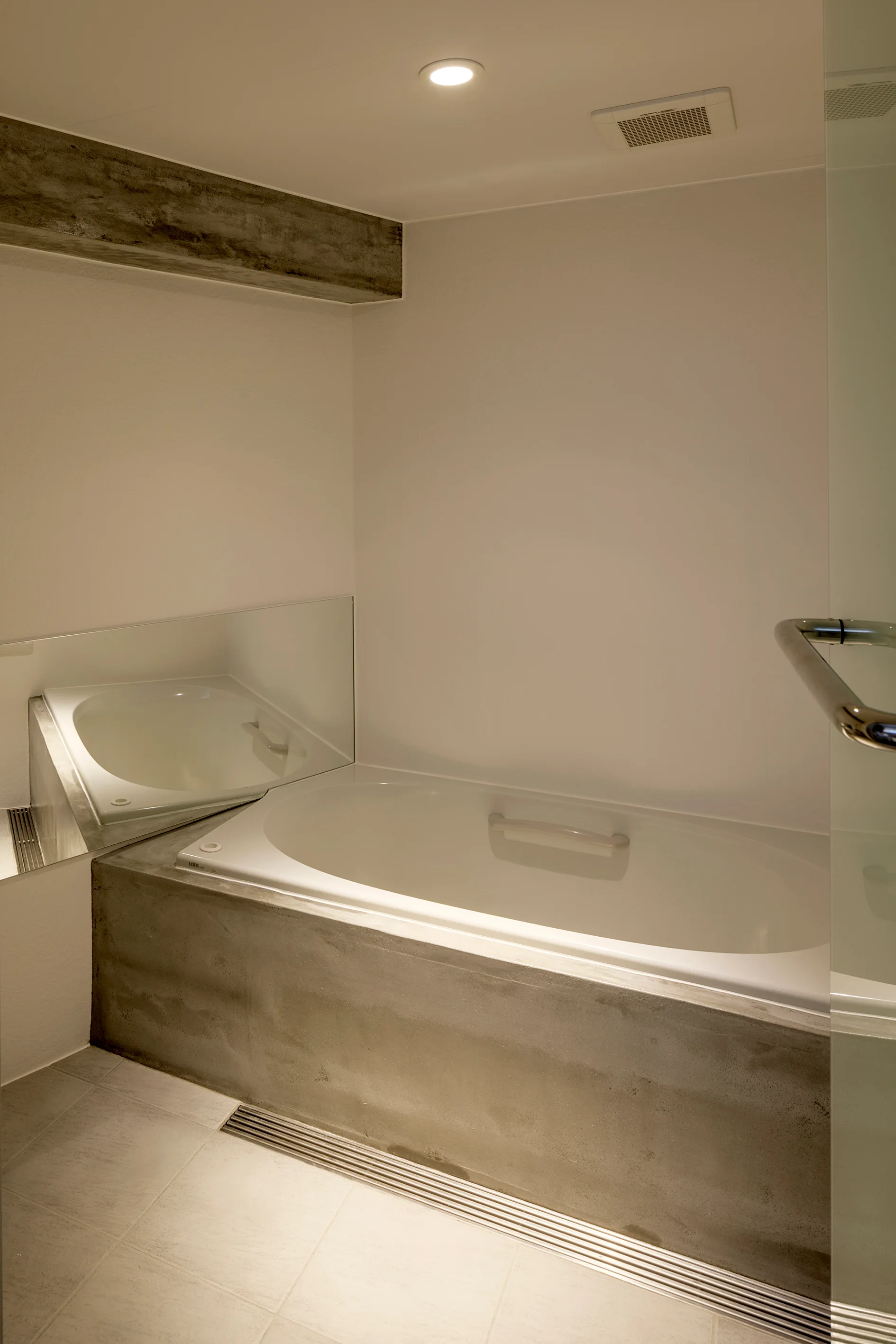 A modern bathroom features a white built-in bathtub with a gray concrete surround, tiled flooring, a glass door, and a mirrored wall panel.