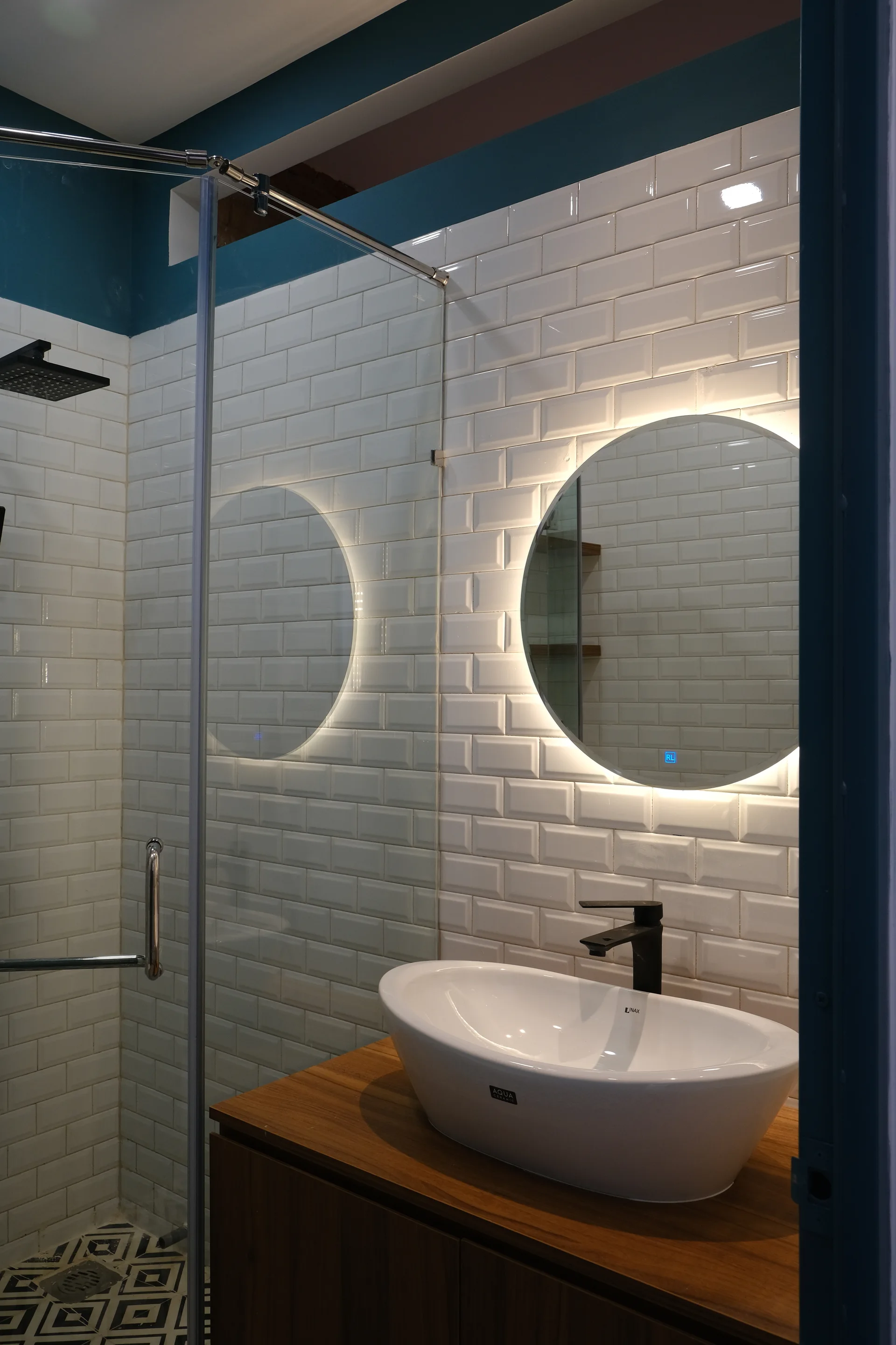 A modern bathroom features a round backlit mirror above a white vessel sink on a wood vanity, set against white glossy subway tile walls.