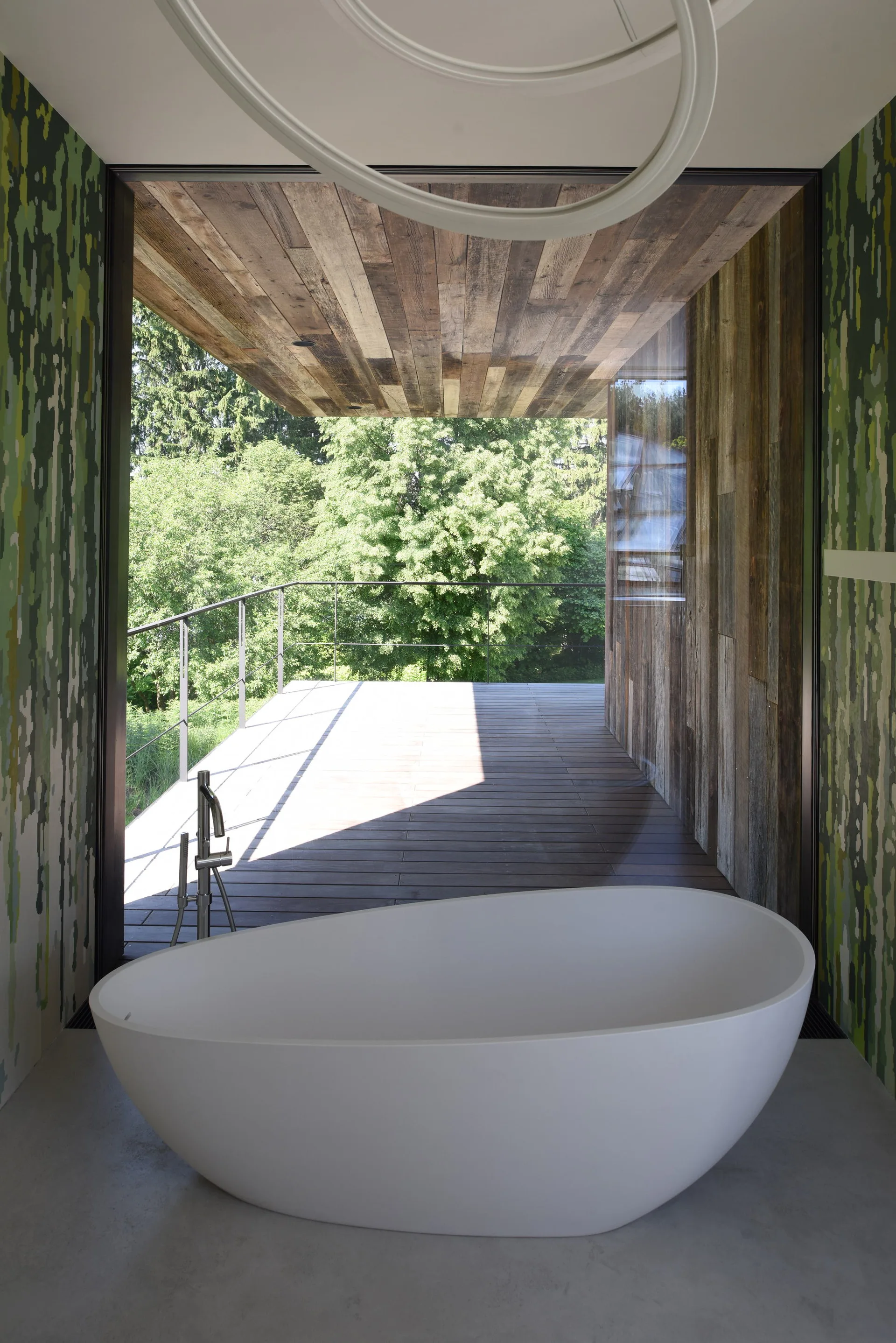 A white freestanding oval bathtub sits on a gray floor in front of a glass wall opening to a wood deck and forest view.