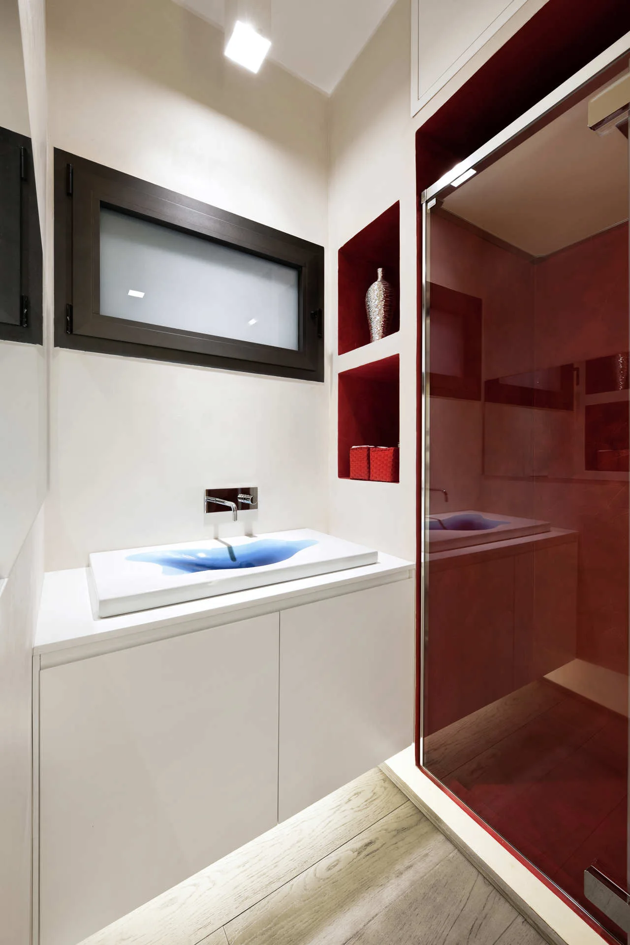 A modern white bathroom with a floating vanity, a unique blue-accented sink, recessed red shelving, and a glass shower.