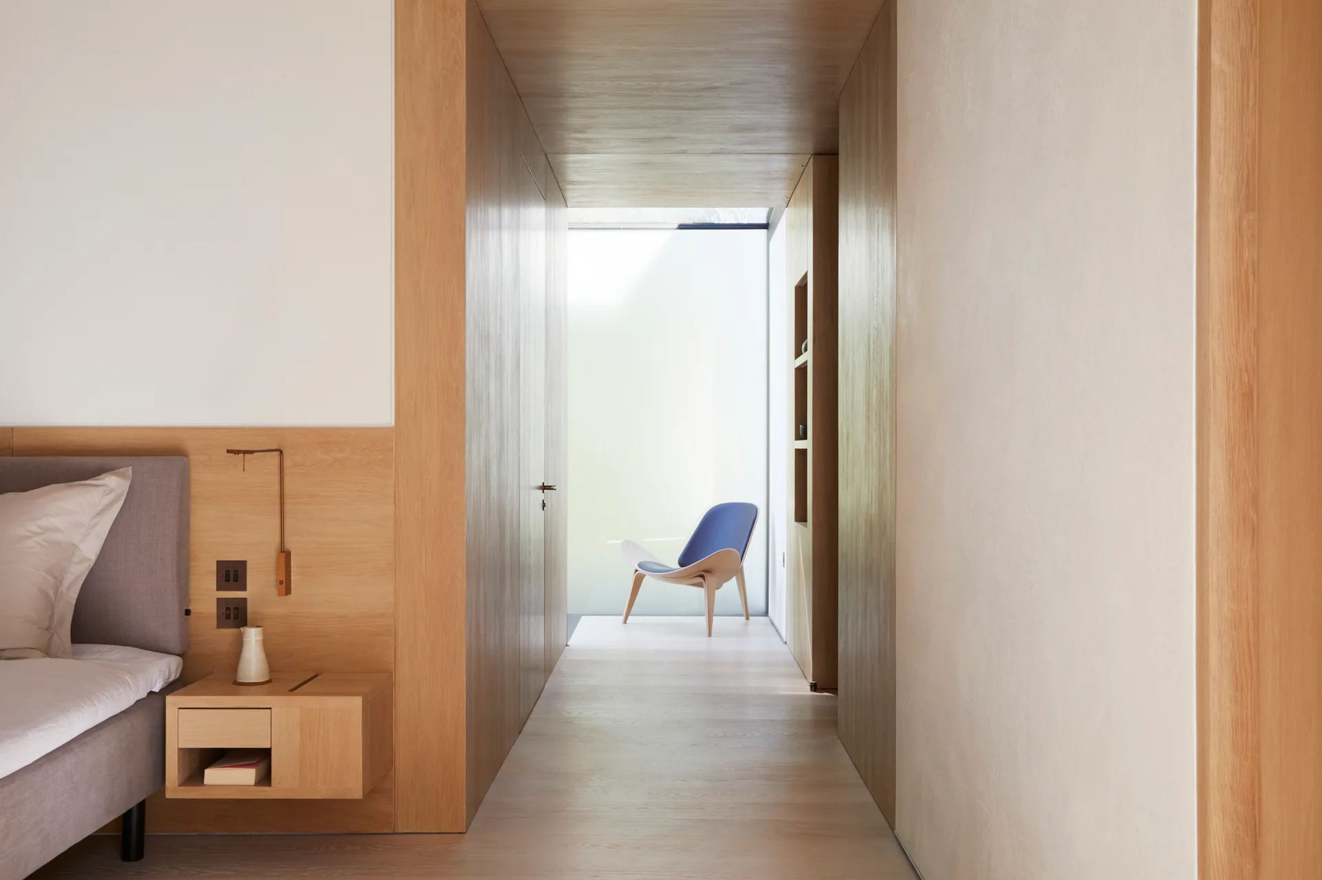 A minimalist bedroom with a floating wood nightstand next to an oak-paneled hallway leading to a blue chair illuminated by natural light.