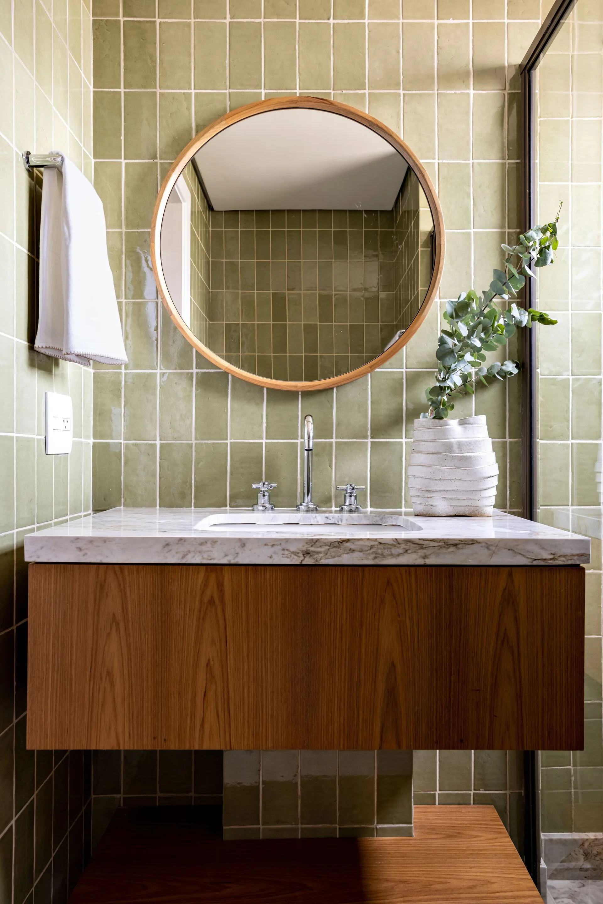 A modern bathroom vanity featuring a floating wood cabinet, marble countertop, chrome faucet, and a round wood-framed mirror against green square wall tiles.