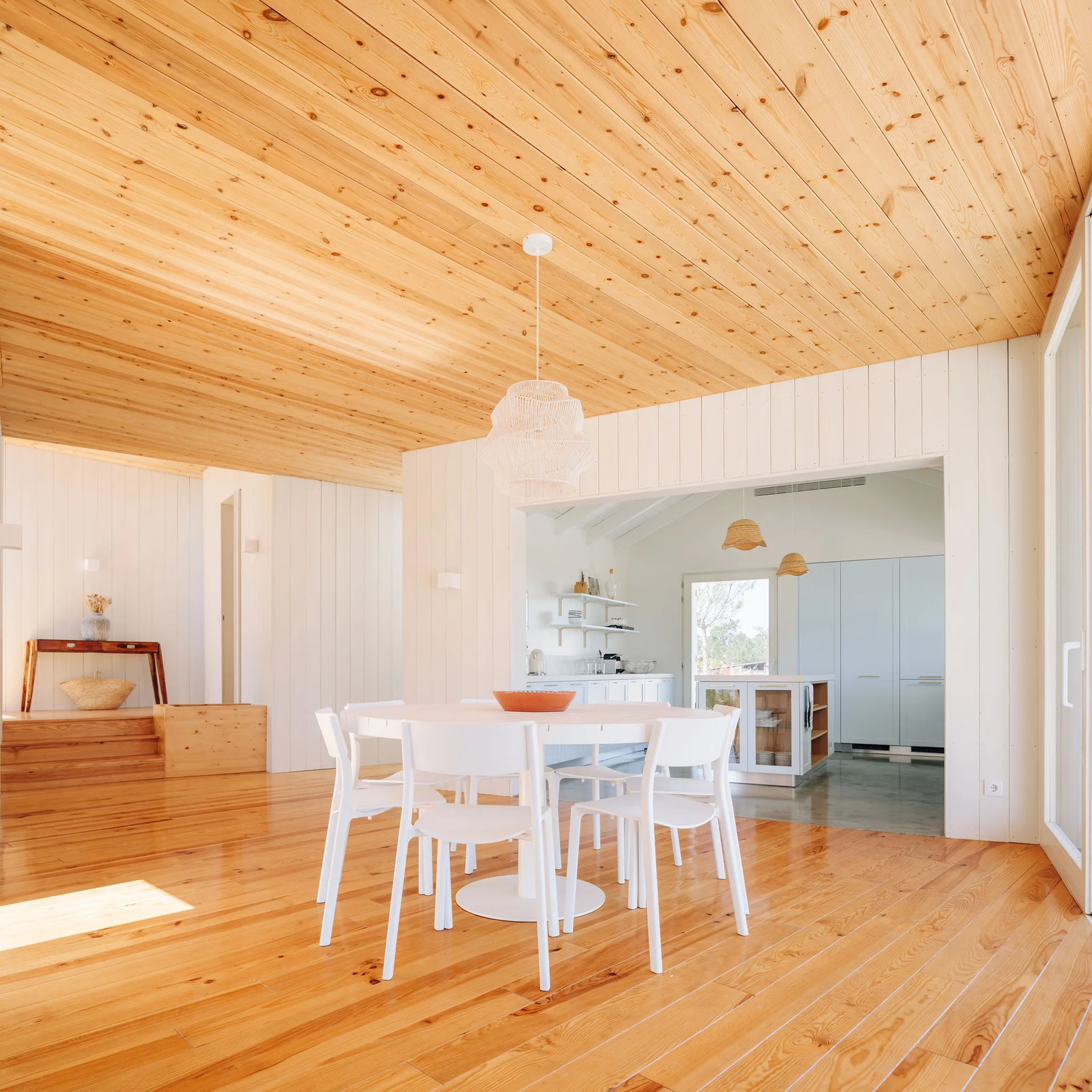 A bright dining room with warm pine flooring and ceiling features a round white dining table, matching white chairs, and a white woven pendant light.
