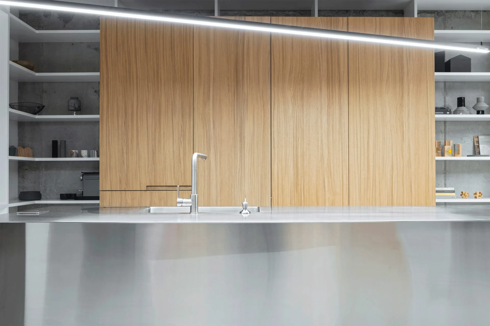 A modern kitchen features a stainless steel island with a sink, backed by wood cabinets, open white shelving, and a raw concrete wall.