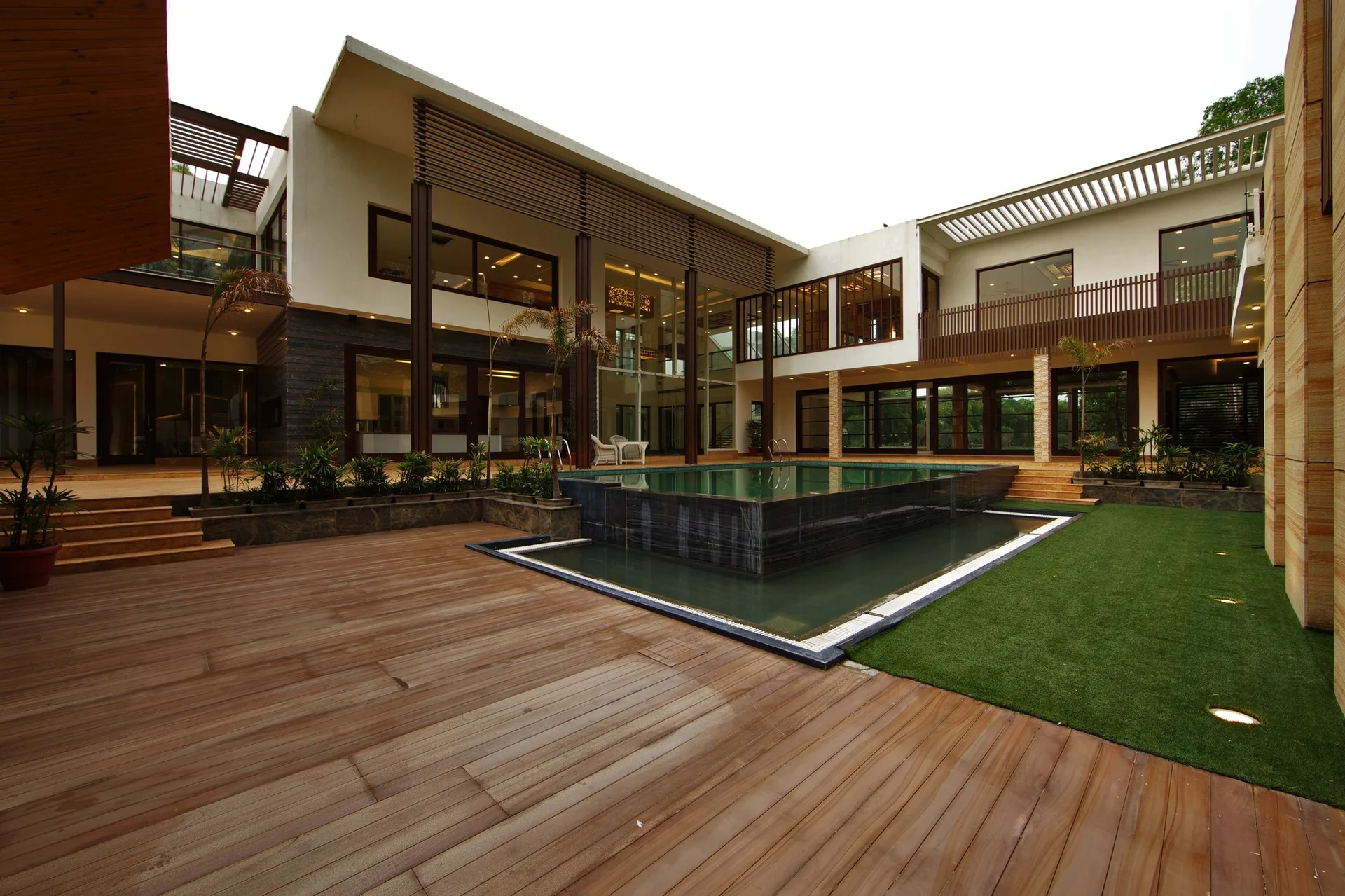 A modern residential courtyard features a raised central swimming pool, expansive wood plank decking, a patch of green grass, and a multi-story home.